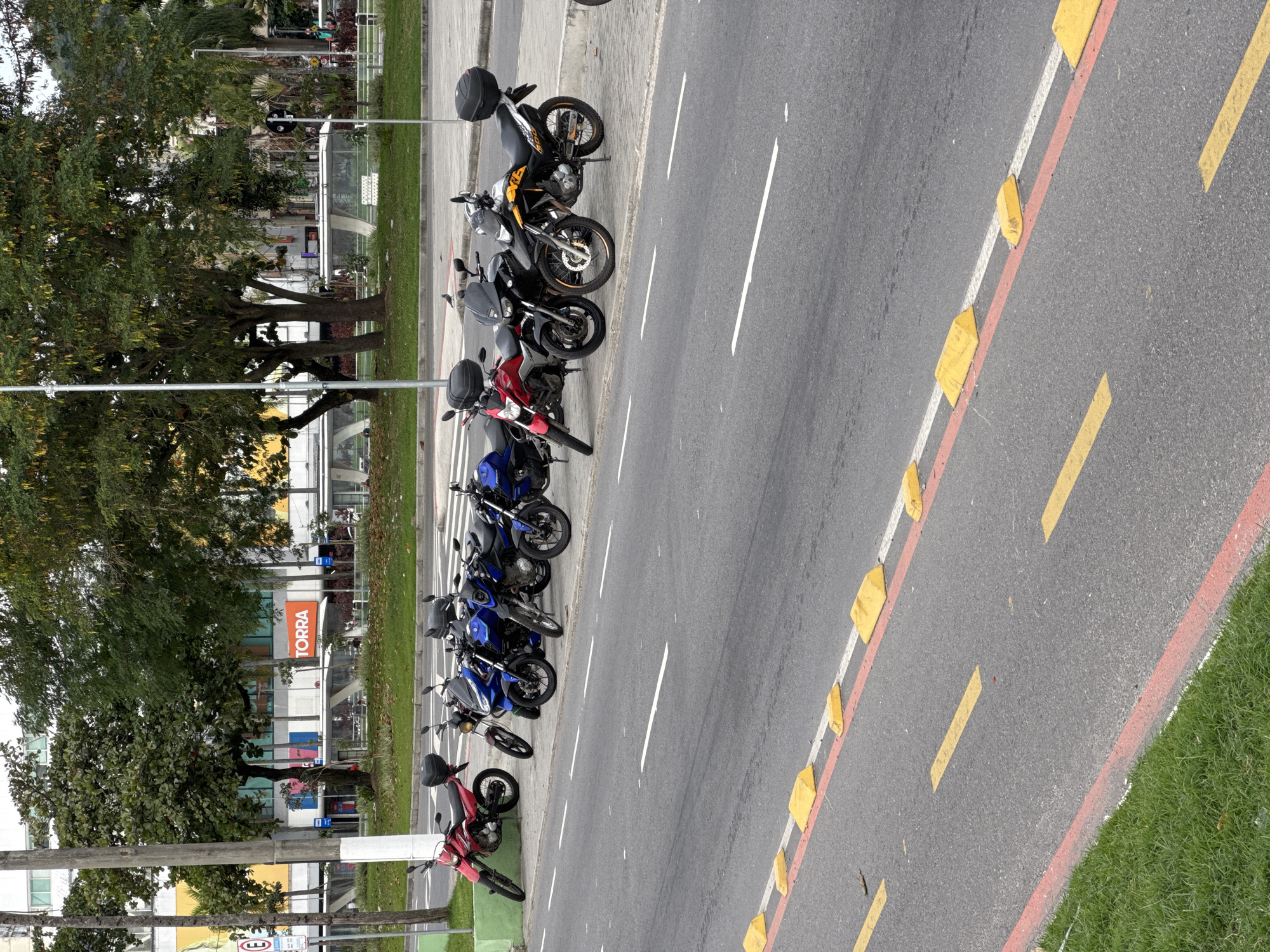 The image captures a street scene in Niterói, Brazil, with a row of parked motorcycles lining the sidewalk. In the background, a white bus is visible, suggesting public transportation is operational. The architecture of a building with "TORRA" prominently displayed in an orange sign indicates a commercial establishment. Trees are present, providing shade and a natural element to the urban landscape. The road is marked with yellow and white lines, and yellow traffic bumps are strategically placed on the road. The overall impression is a typical day in a Brazilian city, with everyday objects and infrastructure.