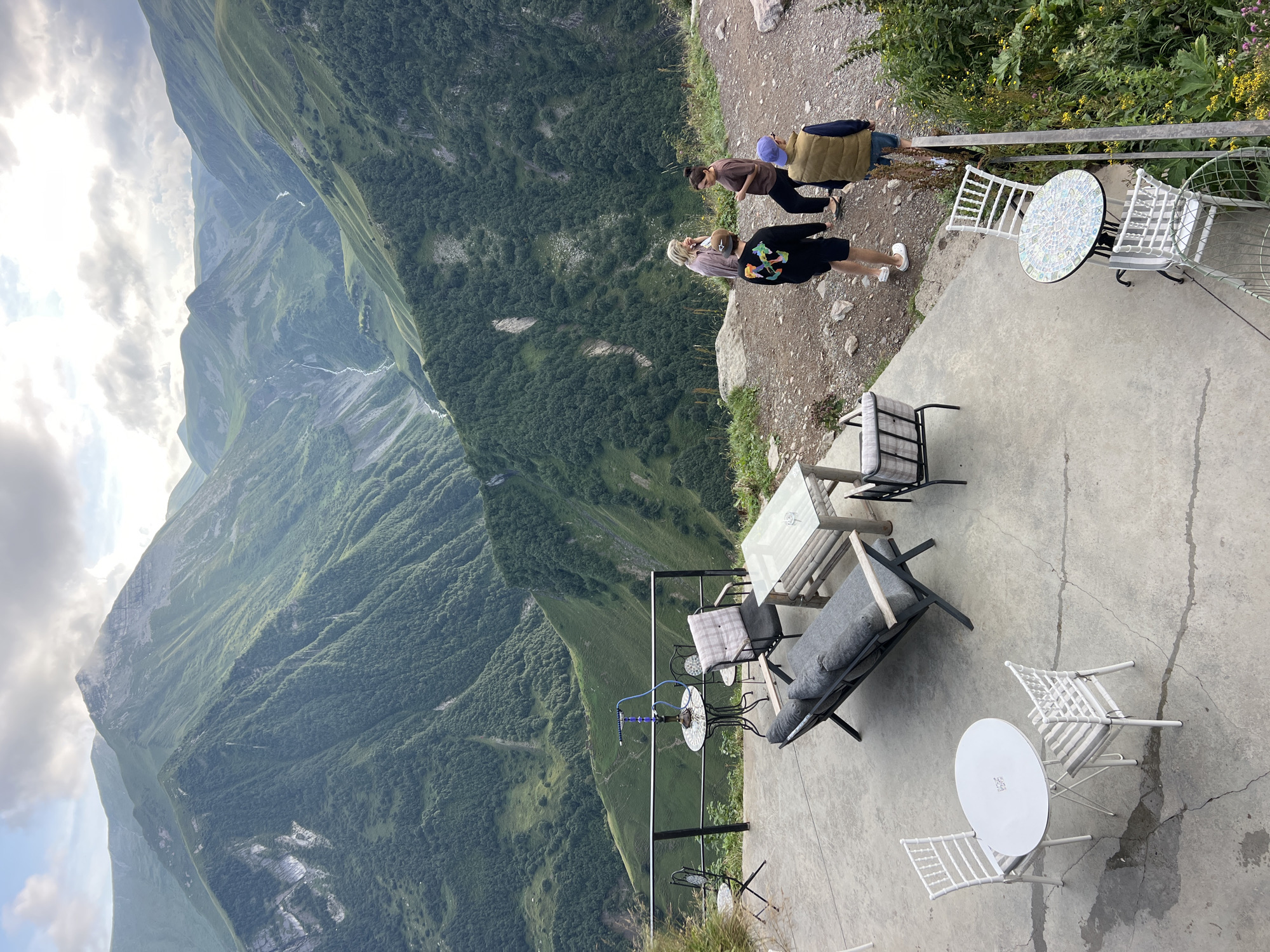 This is a medium-shot, eye-level, outdoor photograph taken at a cafe overlooking a vast mountainous landscape in Gudauri, Georgia. In the foreground, a concrete patio is furnished with several tables and chairs. To the left, a white round table with two white chairs is visible. Further back, a gray sofa with cushions sits next to a rectangular table made of wood and glass. Another smaller table with a mosaic top and two white chairs is located to the right. A hookah is placed on a small table near a railing.
In the middle ground, four individuals are standing and conversing on a gravel path. Two women, one with blonde hair wearing a pink top, and another with dark hair wearing a brown top, are facing each other. Two men, one wearing a black hoodie and shorts, and the other in a tan vest and shorts, are standing with their backs to the camera.
The background is dominated by immense, lush green mountains stretching into the distance. The slopes are covered with dense vegetation and trees, and deep valleys are carved into the terrain. A river or stream can be seen winding through one of the valleys. The sky is partly cloudy with a mix of light and dark clouds. The overall impression is one of natural beauty and a serene, elevated outdoor seating area.