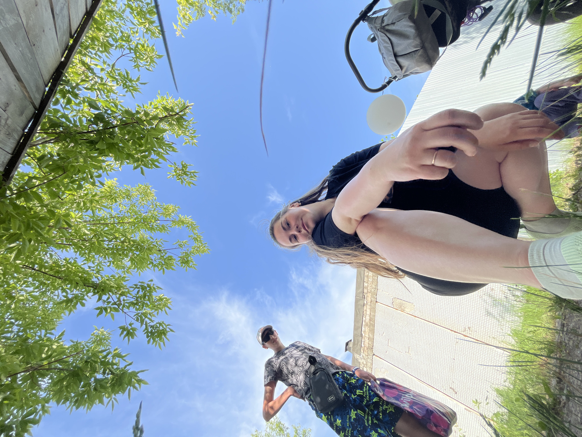The image, taken from a low angle, depicts two people outdoors in what appears to be a partially urban environment.
A woman is positioned closer to the camera, squatting and looking down at the viewer. She has long blonde hair and is wearing a black top. A silver ring is visible on one of her fingers. Her legs and feet are in the frame.
A second individual, a man, is seen further in the background and appears smaller due to perspective. He is wearing a patterned t-shirt, sunglasses, and patterned blue shorts. He has a black bag slung across his shoulder and is holding a pink patterned bag.
Above the individuals, the sky is visible with scattered clouds and a faint trail. To the left, there is foliage with green leaves, suggesting trees. To the right, there is a white surface, possibly a building wall, with some vegetation growing along the base.
The location is Chaikovsky, Russia.