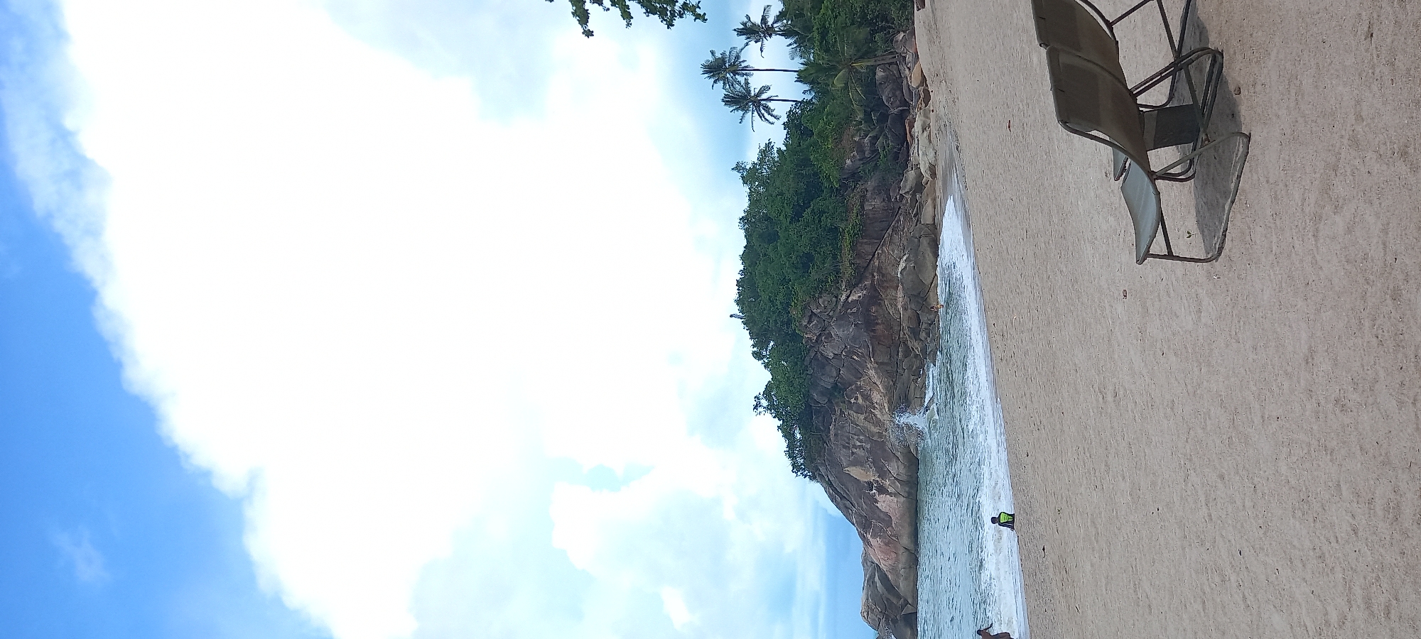 The image shows a coastal scene with a sandy beach, rocky cliffs covered in vegetation, and the ocean. In the upper part of the frame, there are a few palm trees visible. A set of beach chairs is positioned on the sand. Two people are present in the water. The sky is partly cloudy. The image is taken at the location of Baan Thong Nang, Thailand.