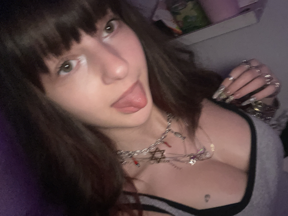 A woman with dark brown hair and bangs sticks her tongue out at the camera. She wears multiple silver necklaces, one with a Star of David pendant. She is dressed in a gray tank top with black trim. Her fingernails are long and decorated with silver rings on each finger. She is indoors and there appears to be a shelf with items above her.