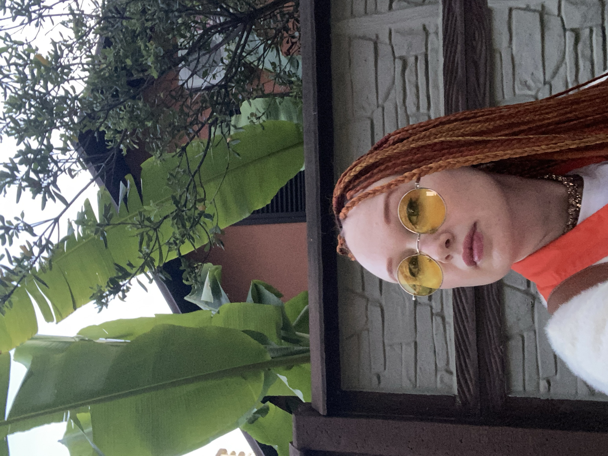 The image presents a close-up, head-and-shoulders portrait of a person outdoors, viewed from a slightly low angle. The subject, appearing to be a young woman, has fair skin and long, reddish-brown hair styled in distinct braids. She is wearing round, yellow-tinted sunglasses, which obscure her eyes, and has light pink lipstick on her lips. Her attire includes a vibrant orange high collar and a white or cream-colored fluffy garment, possibly a jacket or sweater. A subtle gold-toned chain or earring is visible near her neck/ear.
The background is a mix of lush subtropical vegetation and architectural elements. On the left side of the frame and extending upwards, large, vivid green banana leaves and smaller, denser green foliage from other trees or bushes are prominent. Through the gaps in the greenery, a building with an orangish-brown wall and dark window frames or louvers can be seen. To the right of the subject, a textured wall, possibly made of light-colored stone or concrete blocks, forms the backdrop. A dark, vertical wooden or metallic beam or frame separates the subject and the textured wall from the left side's abundant foliage.
The lighting is bright and even, indicating daytime, possibly under an overcast sky or soft natural light. The tropical plants are a strong cue for a warm climate, consistent with a location like Sochi, Russia. The person is looking straight ahead or slightly to the side, appearing to be posing for the photograph with a neutral expression. No text is visible in the image.