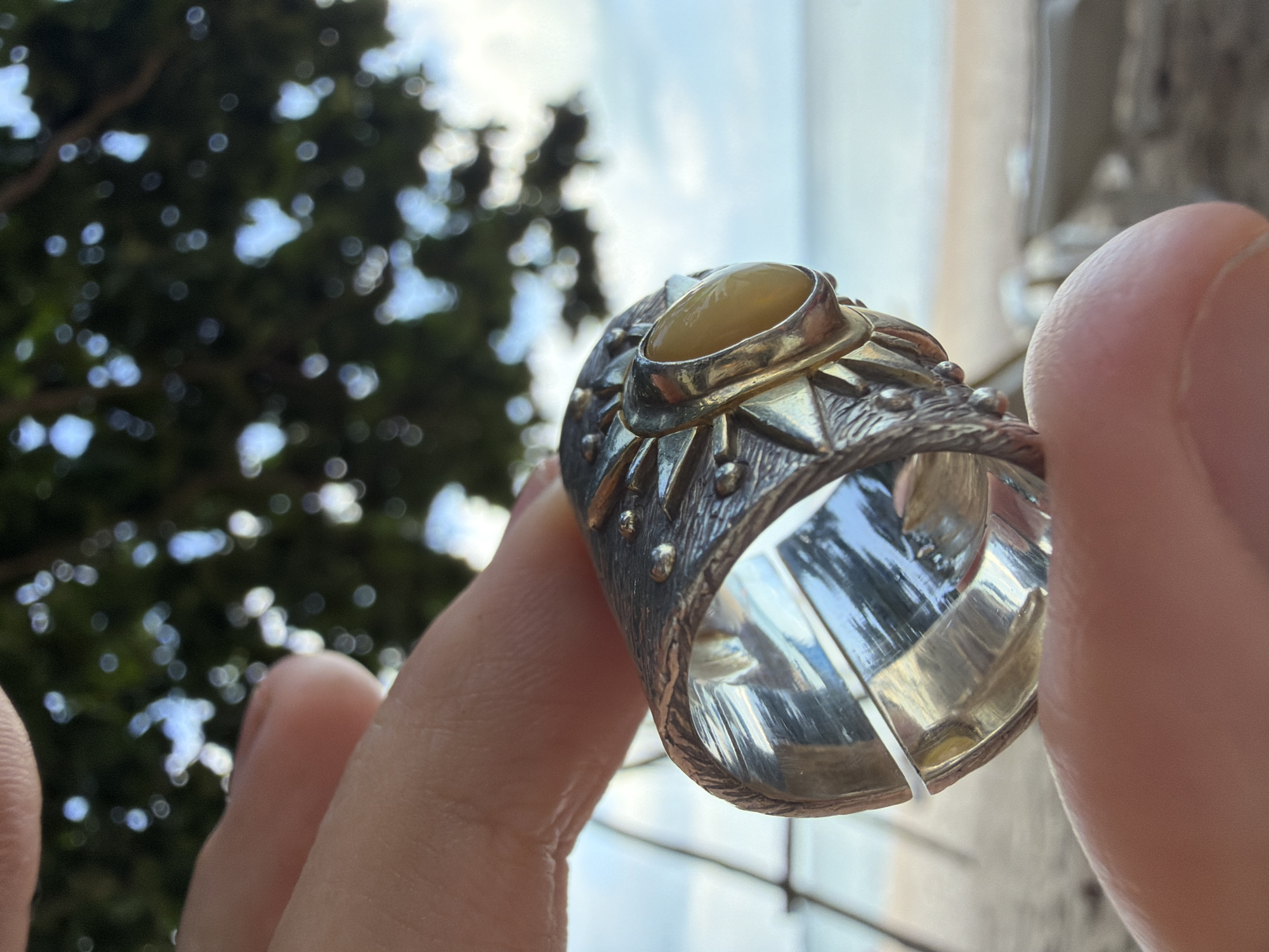 A hand holds a silver ring with an amber stone in the foreground against a blurred background of a beach in Ko Samui District, Thailand. The ring is detailed with a textured band and star-like embellishments. The beach setting is indicated by the sand, the ocean, and a hint of greenery and sky. The focus is on the ring and the hand.