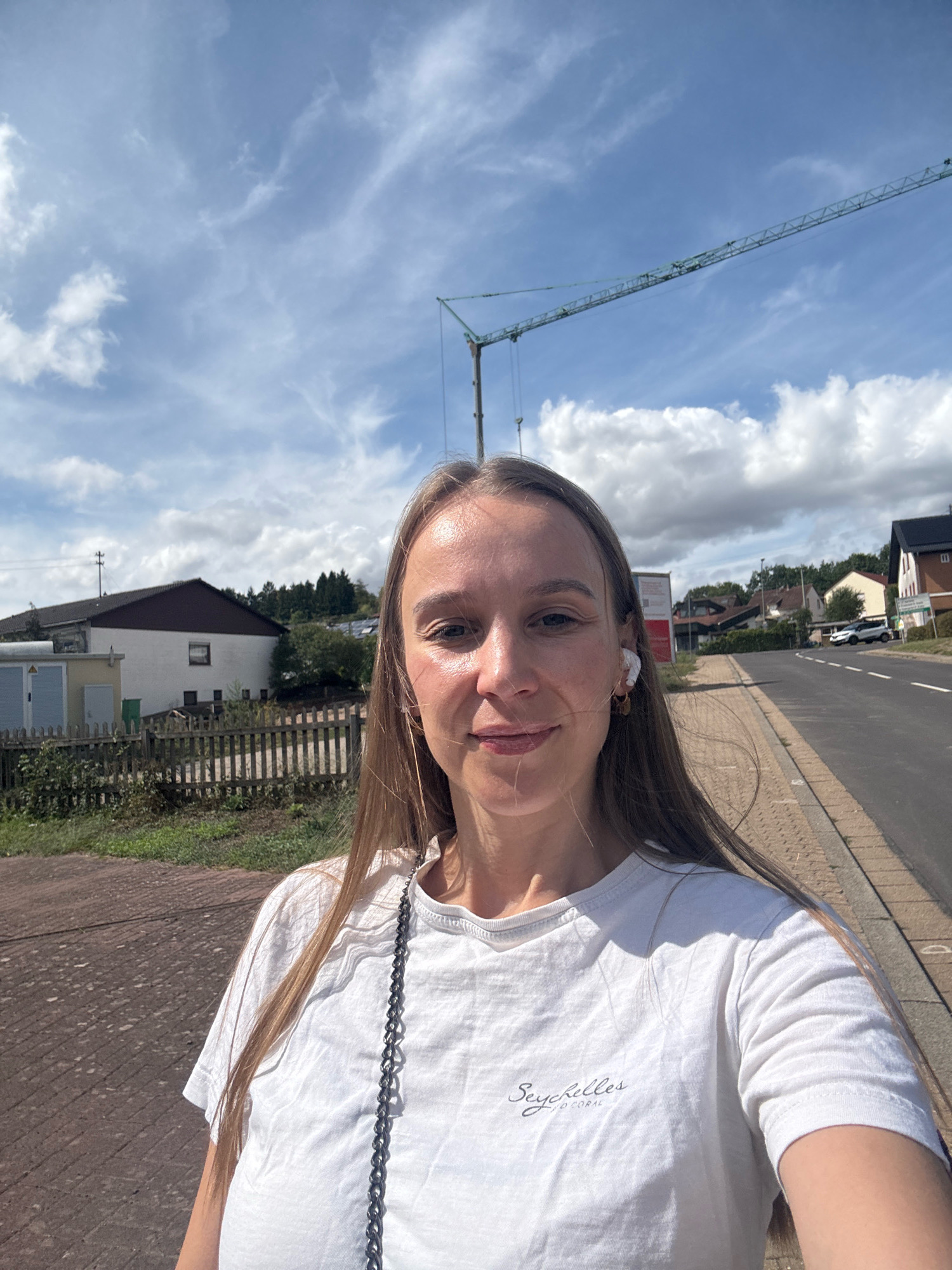 The image shows a young woman with long, light brown hair, wearing a white t-shirt with the inscription "Seychelles" and "O'CORAL" printed on it. She has white earbuds in her ears and is taking a selfie outdoors on a sunny day. In the background, a construction crane is visible against a blue sky with white clouds. To the left of the woman, there are buildings, a wooden fence, and greenery. To her right, a road stretches into the distance, with houses and trees lining the sides. The scene appears to be located in a suburban or semi-rural area of Überherrn, Germany, likely near a construction site given the presence of the crane. The pavement in the foreground suggests she is on a sidewalk or walking path.