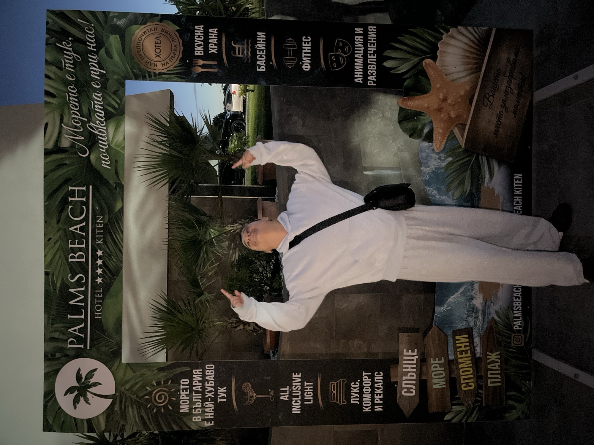 A person stands in front of a photo booth frame that reads "PALMS BEACH HOTEL ★★★★ KITEN". The frame is decorated with tropical leaves and features various text and icons related to a hotel's amenities. On the left side of the frame, text in Bulgarian mentions "The sea in Bulgaria is the most beautiful here". Icons and text describe "ALL INCLUSIVE LIGHT", "LUXURY, COMFORT AND RECREATION", and directional signs pointing to "SUN", "SEA", "MEMORIES", and "BEACH". On the right side, icons and text indicate "DELICIOUS FOOD", "SWIMMING POOLS", "FITNESS", and "ANIMATION AND ENTERTAINMENT". The person is wearing a white hoodie and pants and is looking upwards with their arms raised. Behind the photo booth, palm trees and a building are visible, along with cars in the background. The overall scene suggests a resort or hotel environment in Kiten, Bulgaria, during the evening.