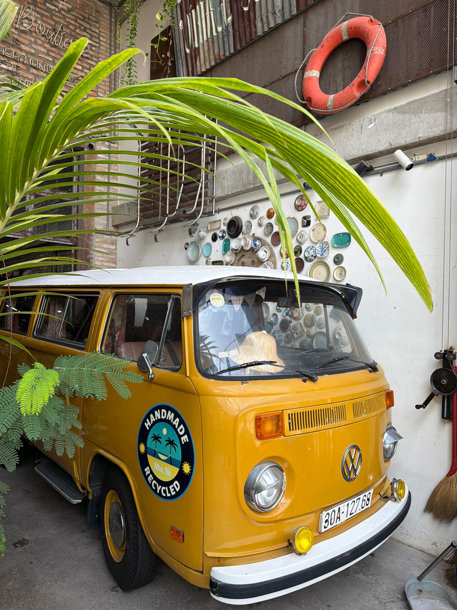 The image depicts a yellow Volkswagen van parked outdoors, likely in the city of Nha Trang, Vietnam, given the Vietnamese text on the circular decal on the van's side. In the foreground, a large, leafy palm frond curves into the frame from the left, partially obscuring the view of the van. The van itself is a vintage model, with a white roof and a prominent circular decal on its passenger side featuring palm trees, a hammock, and the text "HANDMADE NHÀ HÈ RECYCLED." The van has a license plate that reads "30A-127.68".
Behind the van, the wall is adorned with a collection of decorative plates of various sizes and patterns, hung in a somewhat irregular arrangement. To the upper right, a red life preserver with white banding is mounted on the wall. The background includes elements of what appears to be a building facade, with brickwork visible on the left and corrugated metal roofing further up and to the right. The overall scene suggests an outdoor seating or display area, possibly associated with a shop or cafe, given the "Boutique" lettering visible on the brick wall in the upper left corner. The lighting indicates it is daytime.