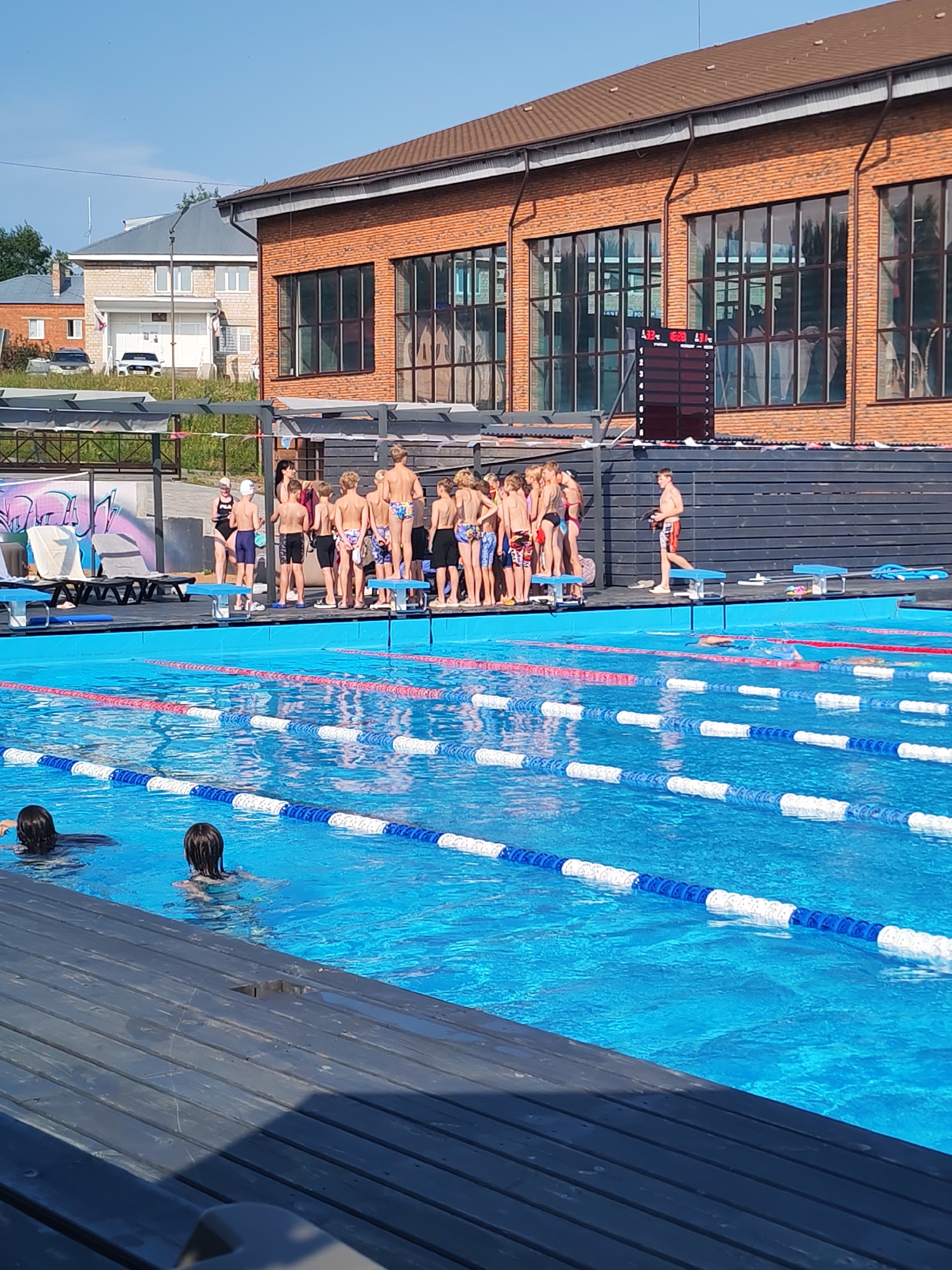An outdoor swimming pool facility in Pirogovo, Russia, is shown on a sunny day. A group of young males, likely swimmers, stand on the deck beside the pool, many in swim trunks. Some are facing the water, others are looking towards a scoreboard. One person is walking on the deck. The pool itself has blue water and lane markers. Two individuals are swimming in the pool. Behind the pool, a brick building with large windows is visible, and further in the background, residential buildings are present. The scene suggests a swimming competition or training session.