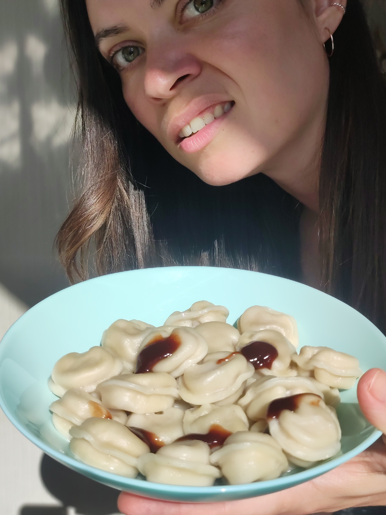 A woman with brown hair and green eyes is holding a light blue bowl filled with pelmeni, small dumplings, topped with a dark sauce, possibly soy sauce. She appears to be indoors, with natural light illuminating the scene. Based on the metadata, this image was taken in Chaikovsky, Russia.