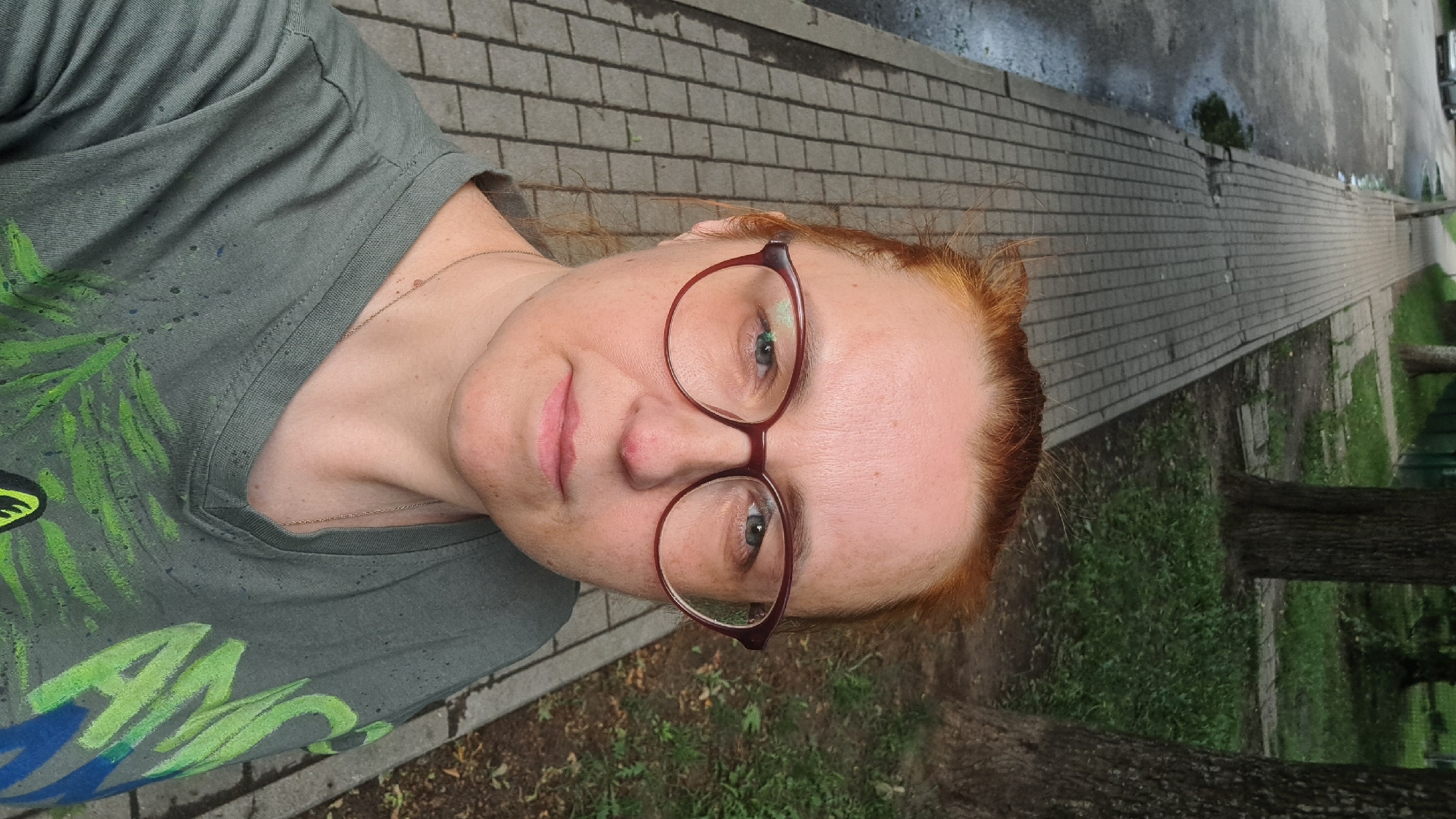 A medium shot, selfie-style photo shows a woman with red hair and glasses looking at the camera. She is standing on a paved sidewalk next to a grassy area with trees. A road with some puddles is visible to her left. The woman is wearing a green t-shirt with a nature-themed print. The scene appears to be an urban park or residential area.