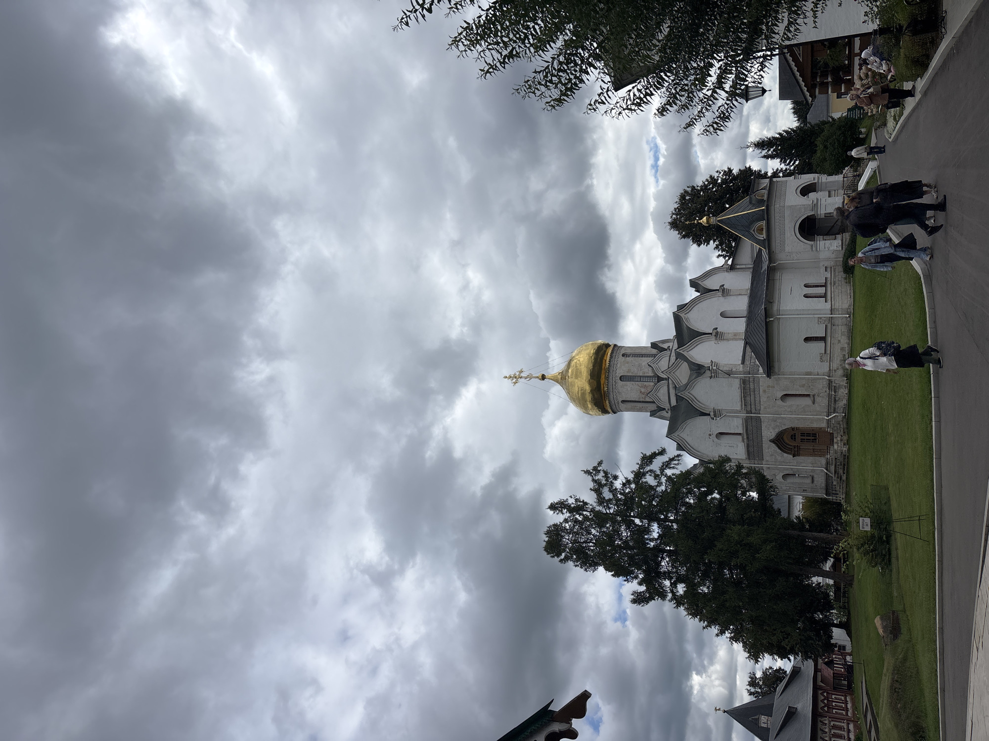 The image depicts the Savvino-Storozhevsky Monastery in Zvenigorod, Russia, under a cloudy sky. A white stone church with a prominent golden dome and a cross is the central focus. To the left of the main church, a smaller, dark-roofed structure with a spire is visible, and further left, part of a red building with ornate trim. In front of the main church, a well-maintained lawn is bordered by a paved pathway. Several people are walking on the path and near the entrance to the church. Some are walking towards the church, while others are moving away or congregating near a building to the right. The foliage surrounding the monastery includes several trees, with one large evergreen tree standing to the left of the main church. The overall atmosphere is overcast, suggesting a potentially cool or damp day.