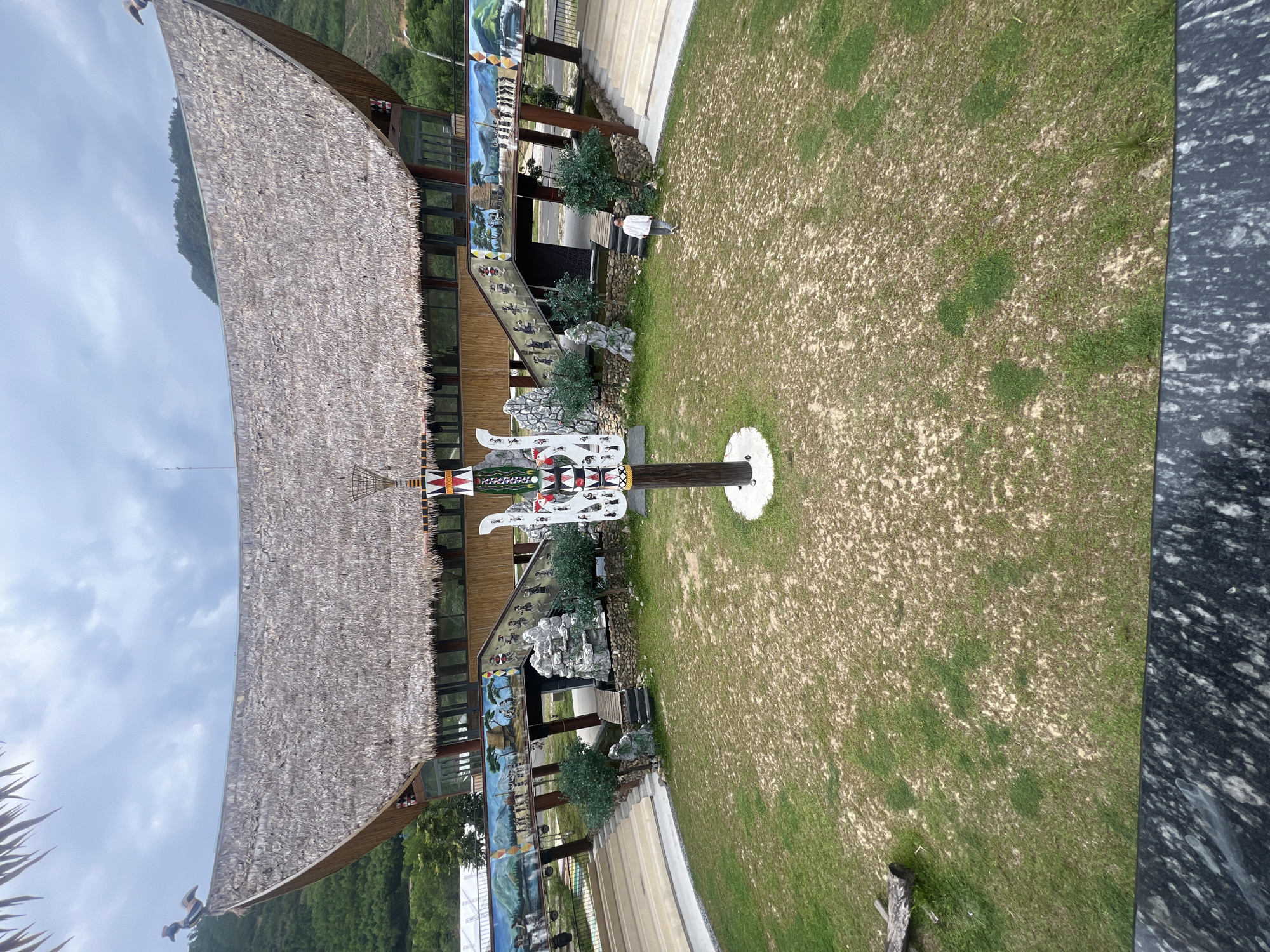 The image captures a wide-angle view of a traditional-style building with a thatched roof, likely part of a cultural park or heritage site. The building features a distinctive curved roofline, adorned with decorative elements that resemble birds on its peaks. A central, intricately carved wooden totem pole stands prominently in the foreground, decorated with geometric patterns and stylized figures.
The building's facade is characterized by wooden architecture with large windows and decorative murals depicting scenes of local life and culture, including people and landscapes. Two staircases with wide steps lead up to the main structure, flanked by small trees and stone landscaping. A single person, dressed in white, is walking away from the viewer towards the building.
The ground in front of the building is a large expanse of grass with patches of dirt, suggesting a somewhat dry or worn area. In the background, lush green hills and trees are visible, indicating a natural, possibly mountainous setting. The sky is overcast with clouds, suggesting a diffused lighting condition. The overall impression is one of a cultural attraction designed to showcase traditional architecture and local heritage. This scene is not in the city of Đà Nẵng, Vietnam.