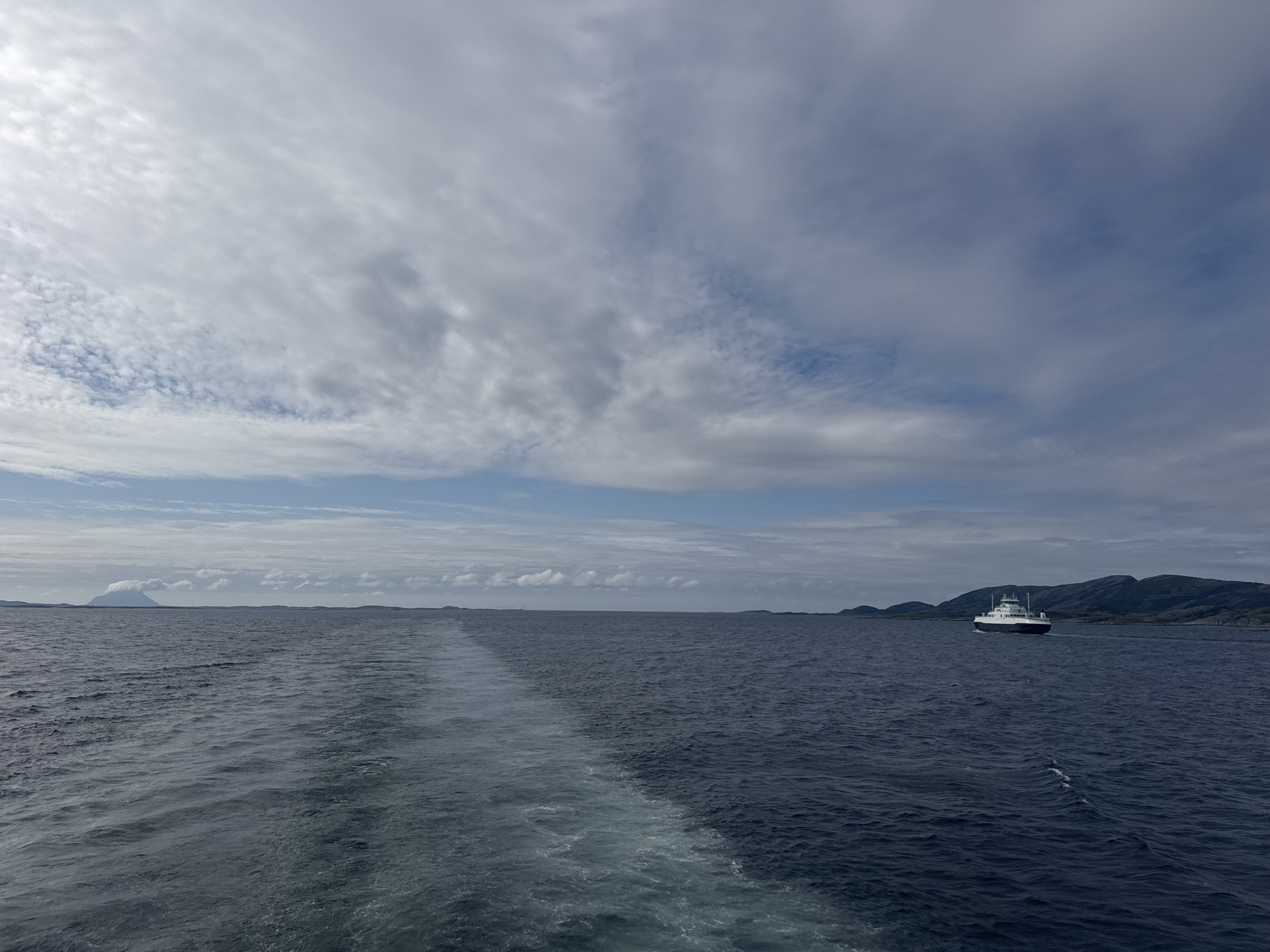 This image depicts a wide shot of the ocean, likely in the vicinity of Stokkvågen, Norway, as suggested by the presence of a ferry. The sea is a deep blue, with visible waves and a prominent white wake trailing from the viewpoint of the camera. In the distance, a white ferry boat with dark trim is visible, navigating the water. Beyond the ferry, a chain of dark, hilly islands or coastal landforms stretches across the horizon under a vast sky filled with a mix of white and grey clouds, with patches of blue sky peeking through. To the far left, a single, distinctly triangular mountain peak rises from the water, capped with clouds. The overall scene is one of a marine journey under a dynamic sky.