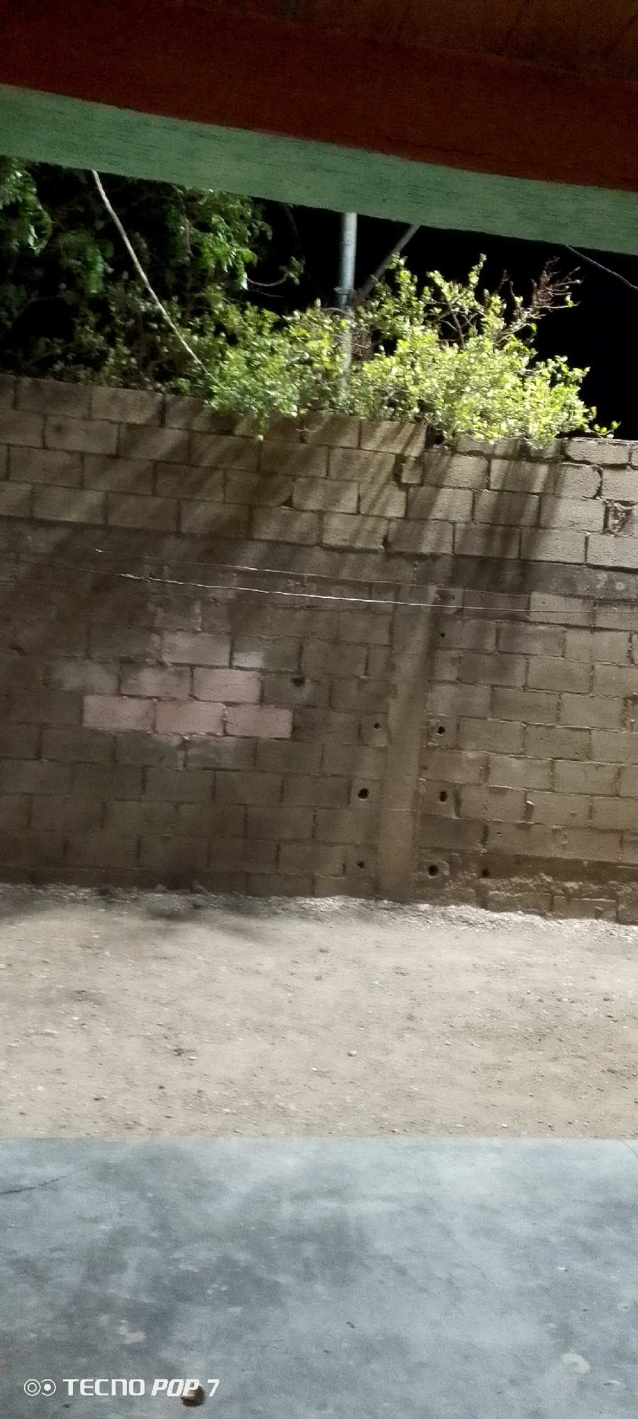 The image depicts a gray brick wall with a patched section of pinkish bricks, topped with green foliage. A dark, possibly wooden, structure is visible above the foliage. A dirt or gravel surface stretches out in front of the wall. The scene appears to be dimly lit, possibly at night. The location in Altagracia, Venezuela, is not explicitly confirmed by the image itself, but provided as external information. No people or actions are visible. The environment appears to be an outdoor area, possibly an alleyway or the edge of a property. A wire stretches horizontally across the wall. The phrase "TECNO POP 7" appears in the lower left corner, likely indicating the device used to capture the image.