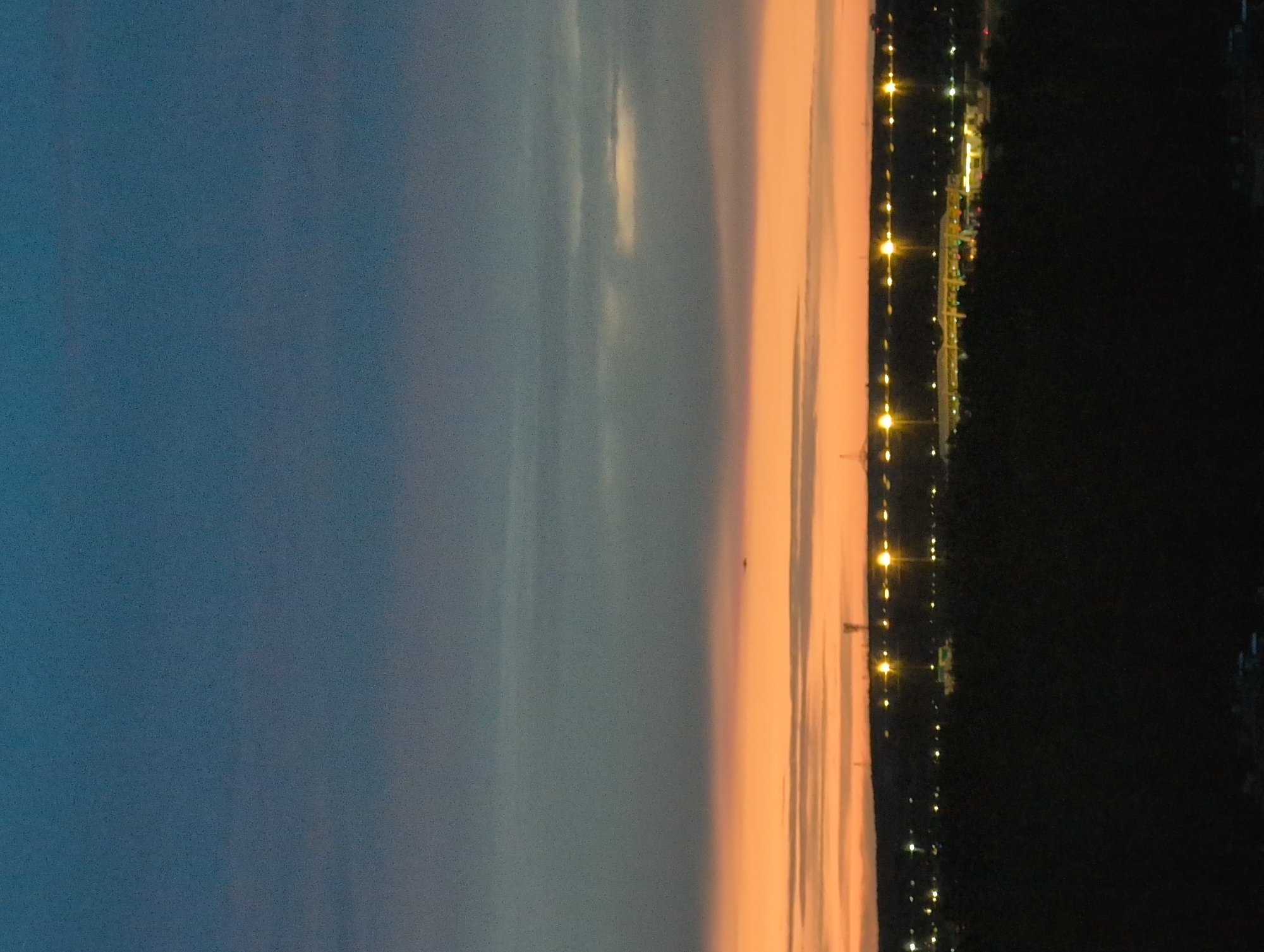 The image depicts a sunset over a city skyline, possibly Saint Petersburg, Russia. The sky transitions from a deep blue at the top to a vibrant orange and pink near the horizon, indicating the sun has recently set or is about to rise. A thin layer of clouds is visible, catching the last rays of light. In the distance, a strip of lights suggests a populated area or a commercial district. Some structures, possibly tall buildings or communication towers, are silhouetted against the twilight. The foreground is dominated by dark, silhouetted trees or foliage, suggesting the viewpoint is elevated and looking over a natural area towards the city. The overall impression is one of a calm, transitional moment between day and night.