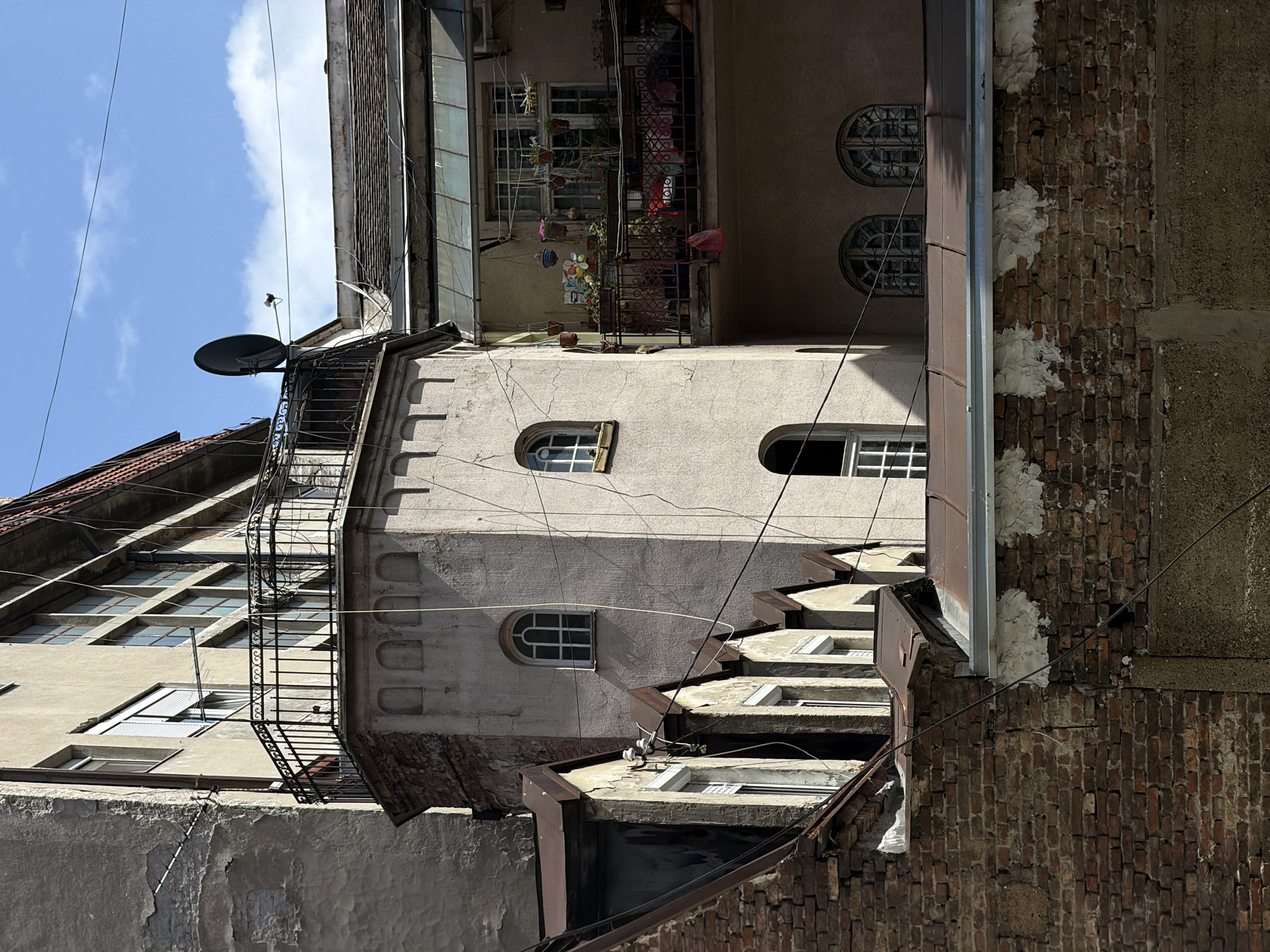 The image captures a cluster of buildings in Belgrade, Serbia, viewed from a low angle, tilted counter-clockwise. The architecture is varied, with a mix of materials including brick, stucco, and stone. A tall, narrow structure with three arched windows is central, featuring an exterior fire escape. Adjacent to this structure is a building with a balcony overflowing with plants and decorations. Satellite dishes are mounted on rooftops. Electrical wires crisscross the scene. The sky is visible in the upper left, displaying patchy blue and white clouds.