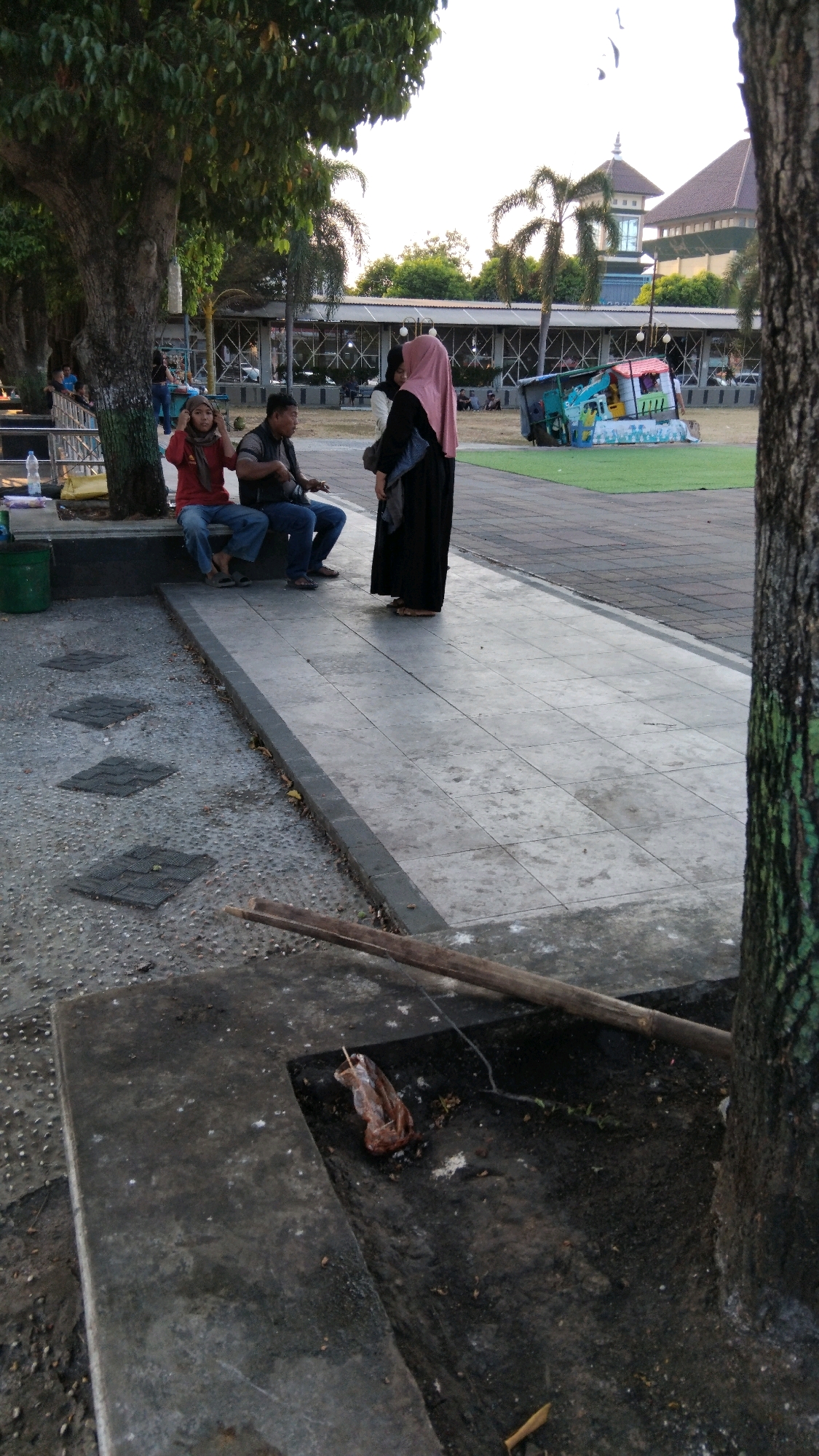 The image depicts an outdoor scene in what appears to be a park or public space in Batang, Indonesia. In the foreground, a concrete area features a large tree trunk on the right and a concrete planter or structure on the left with some debris, including a brown plastic bag and some scattered white particles. A wooden plank lies across the planter.
Further back, a paved walkway leads to a shaded seating area under a large tree. On this seating area, three individuals are visible. A woman in a red top and headscarf sits on the left, facing towards the center. Next to her, a man in a dark vest and blue jeans sits, appearing to be playing a stringed instrument, possibly a guitar. To the right of the man, another individual, a woman in a black abaya and pink headscarf, stands and engages with the seated figures.
Beyond the seating area, the background reveals a paved plaza with a distinctive colorful play structure resembling a truck. Further in the distance, there is a building with an angular roofline and a tower, and rows of palm trees. The sky is bright, suggesting daytime, possibly late afternoon due to the warm light. The overall atmosphere suggests a relaxed, casual gathering in a public urban space.
