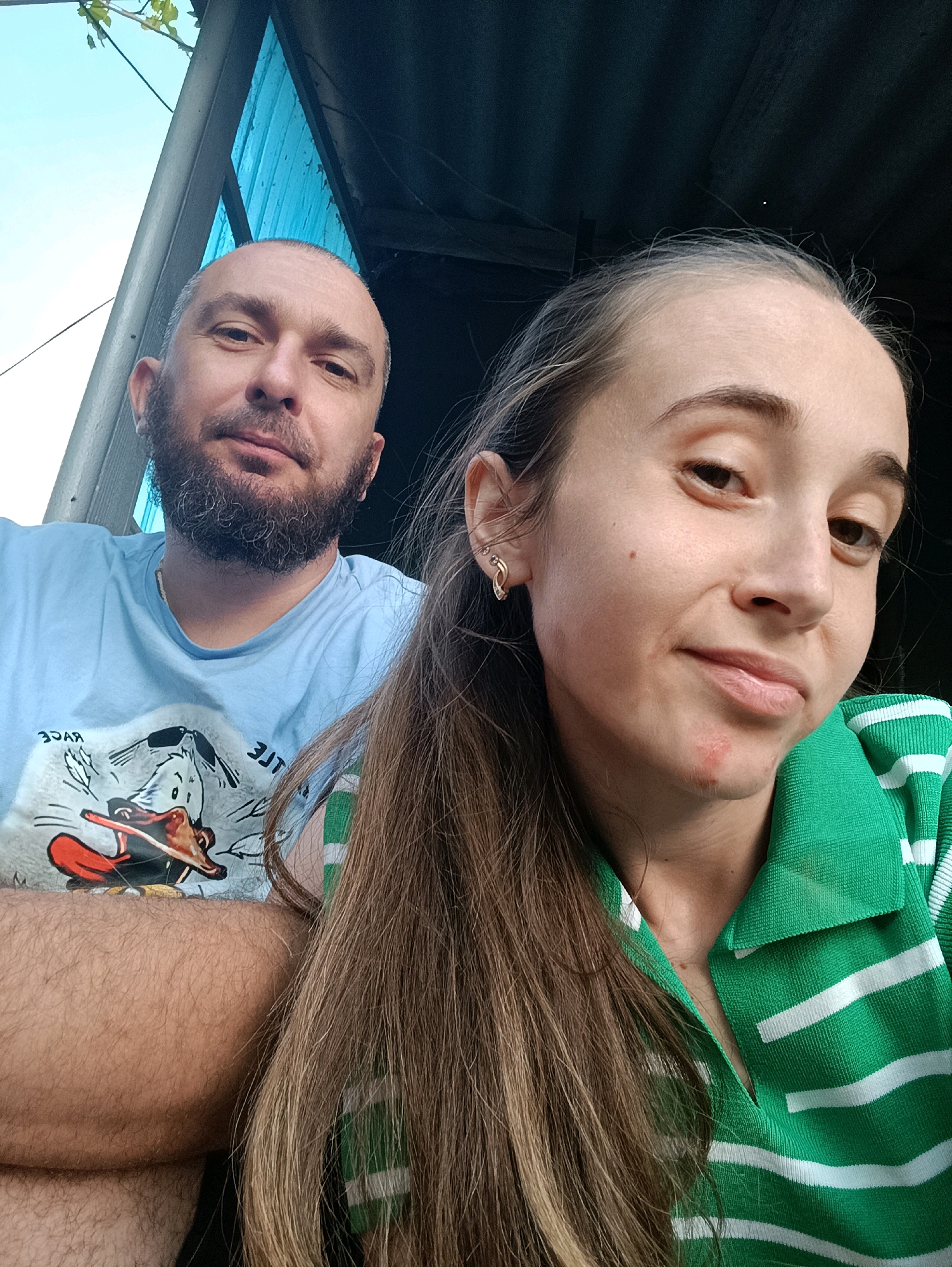 Two individuals, a man and a woman, are pictured in an outdoor setting. The man, with a beard and bald head, wears a light blue t-shirt with a cartoon graphic. He is positioned to the left of the frame, with his arm crossed. The woman, with long brown hair, is on the right, looking towards the camera. She has a gold hoop earring in her left ear and is wearing a green and white striped polo shirt. A red mark is visible on her chin. The background suggests an outdoor structure with a dark roof and a blue wall, possibly in a rural or suburban area. There is greenery visible at the top left of the image. The location is identified as Vypasne, Ukraine.