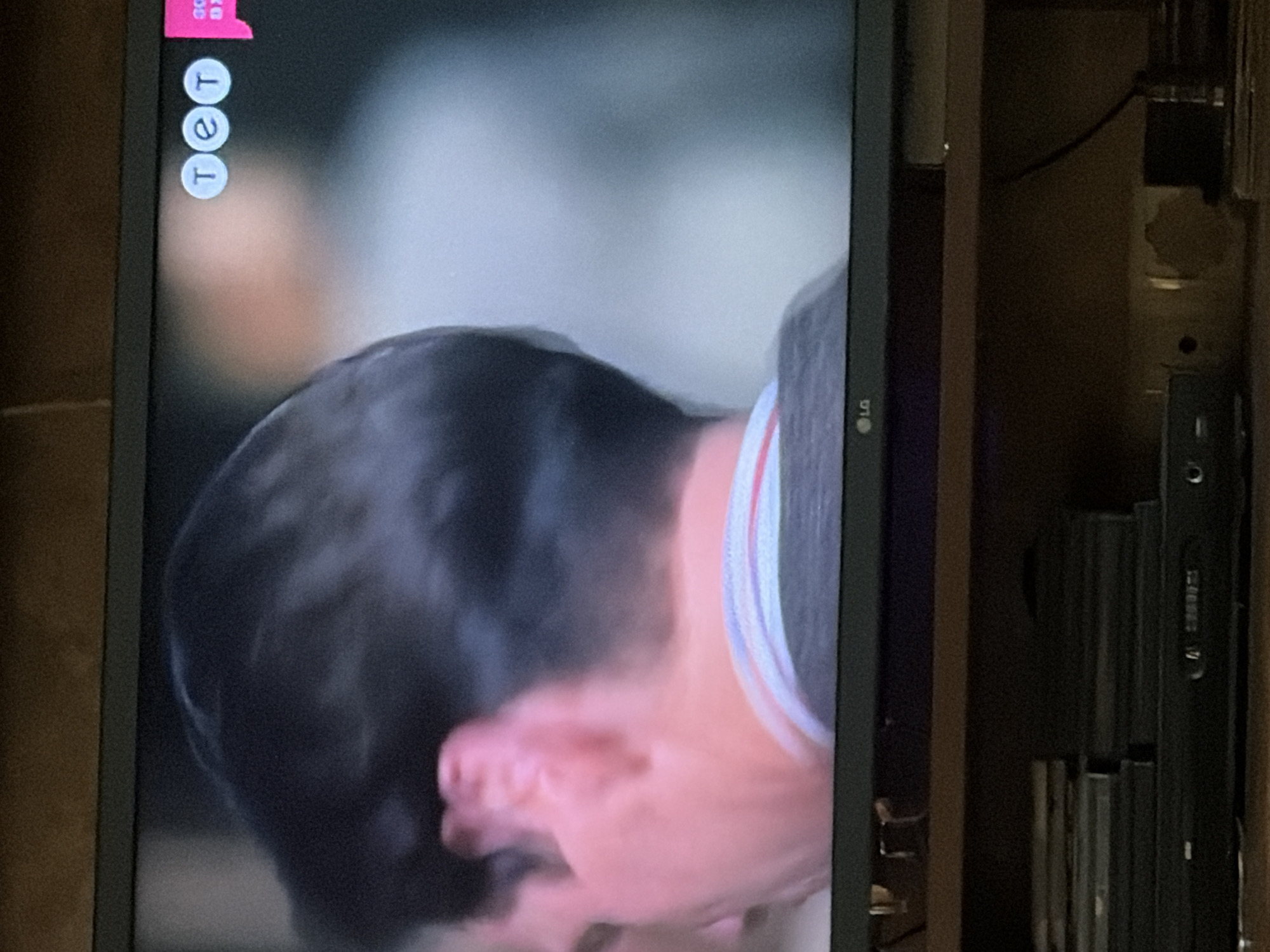 The image captures a close-up view of a television screen displaying a person's head and shoulder, with a visible LG logo at the bottom center of the screen. The background of the television shows a blurred, light-colored scene, possibly an interior setting, with three circular logos in the upper right corner reading "TET". Adjacent to these logos is a pink rectangular graphic. The television is placed on a wooden shelf, beneath which is a black DVD player and a stack of what appear to be CD or DVD cases. The scene is dimly lit, suggesting it is indoors. The location in the city of Osolynka, Ukraine, is not discernible from the image itself.