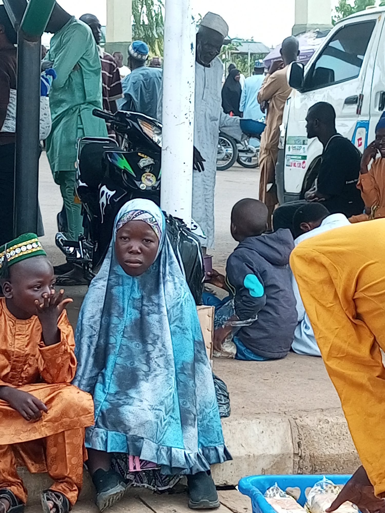In Gombe, Nigeria, a group of people are gathered outdoors, possibly at a market or roadside. In the foreground, a young girl wearing a light blue headscarf and dress sits facing the camera, her expression solemn. Beside her to the left, a young boy in an orange outfit and a green, gold-embroidered cap also sits, his hand near his mouth. Behind them, several men are visible. One man in a green robe stands to the left, while others in various traditional attire are scattered throughout the scene. A motorcycle is parked behind the children, and a white van is to the right. The ground appears to be concrete or paved. A blue plastic container with some items in it is in the lower right corner of the frame. The overall impression is of a bustling public space with individuals engaged in everyday activities.
