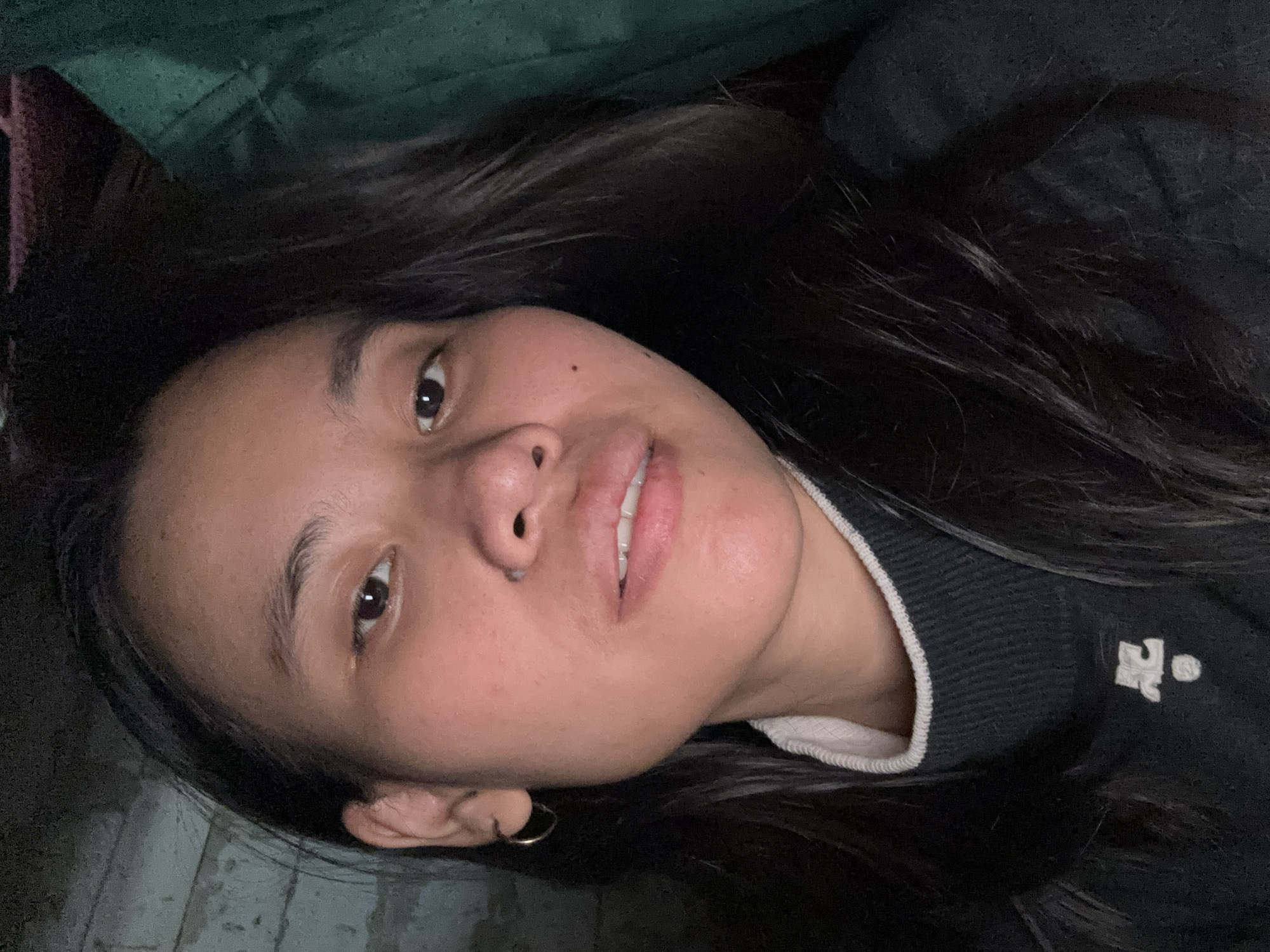 A close-up selfie of a young woman with dark hair is shown. She is wearing a dark gray sweater with a white trim around the neck and a small white emblem on the left side of the chest. Her left ear is visible and adorned with a small hoop earring. The background is dark and appears to be an indoor setting with a brick wall on the left and a draped green fabric on the right. The lighting is dim. No specific location in East Jakarta, Indonesia is indicated by the image.