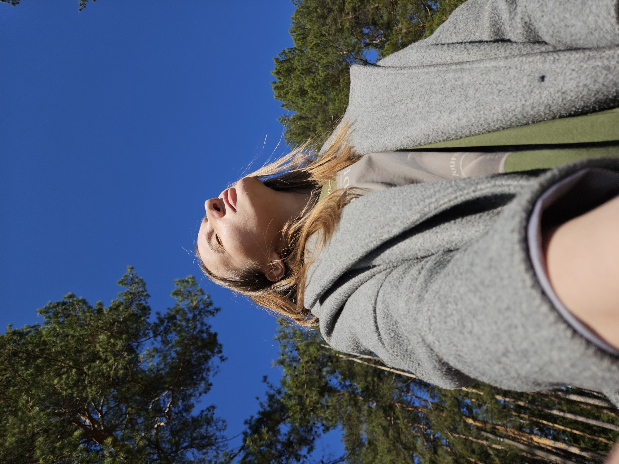 The image shows a woman from a low-angle perspective, gazing upwards. She is wearing a grey coat and a green shirt. The background consists of a bright blue sky and treetops. The image was taken outdoors in a wooded area, likely in the city of Chaikovsky, Russia. The image is oriented with the subject looking upwards towards the sky, framed by the trees.