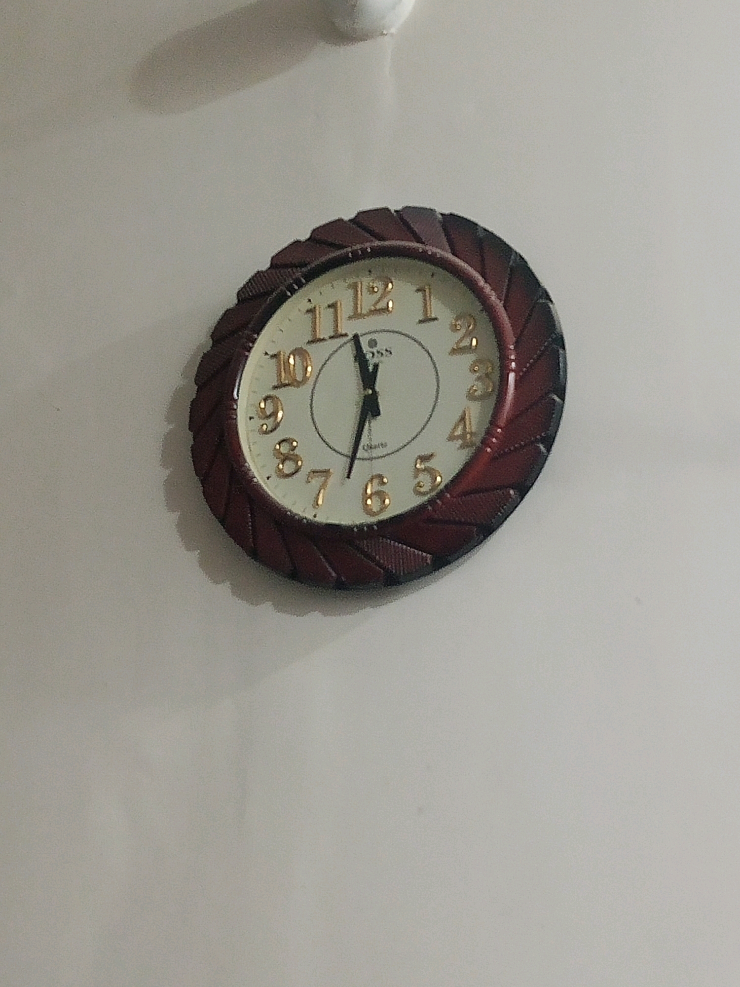 A dark brown decorative analog wall clock is mounted on a light-colored wall. The clock face is white with gold numerals and hands. The hour hand is pointing at approximately 10, and the minute hand is pointing at approximately 5. The clock has a textured, dark brown bezel. The wall appears to be indoors, and a shadow from a light fixture or an object is visible on the wall above the clock. The image is taken from a slightly angled perspective. The location of Bhedian - Nai Basti, Pakistan is provided, but there is no visual information in the image to corroborate or deny this specific location.
