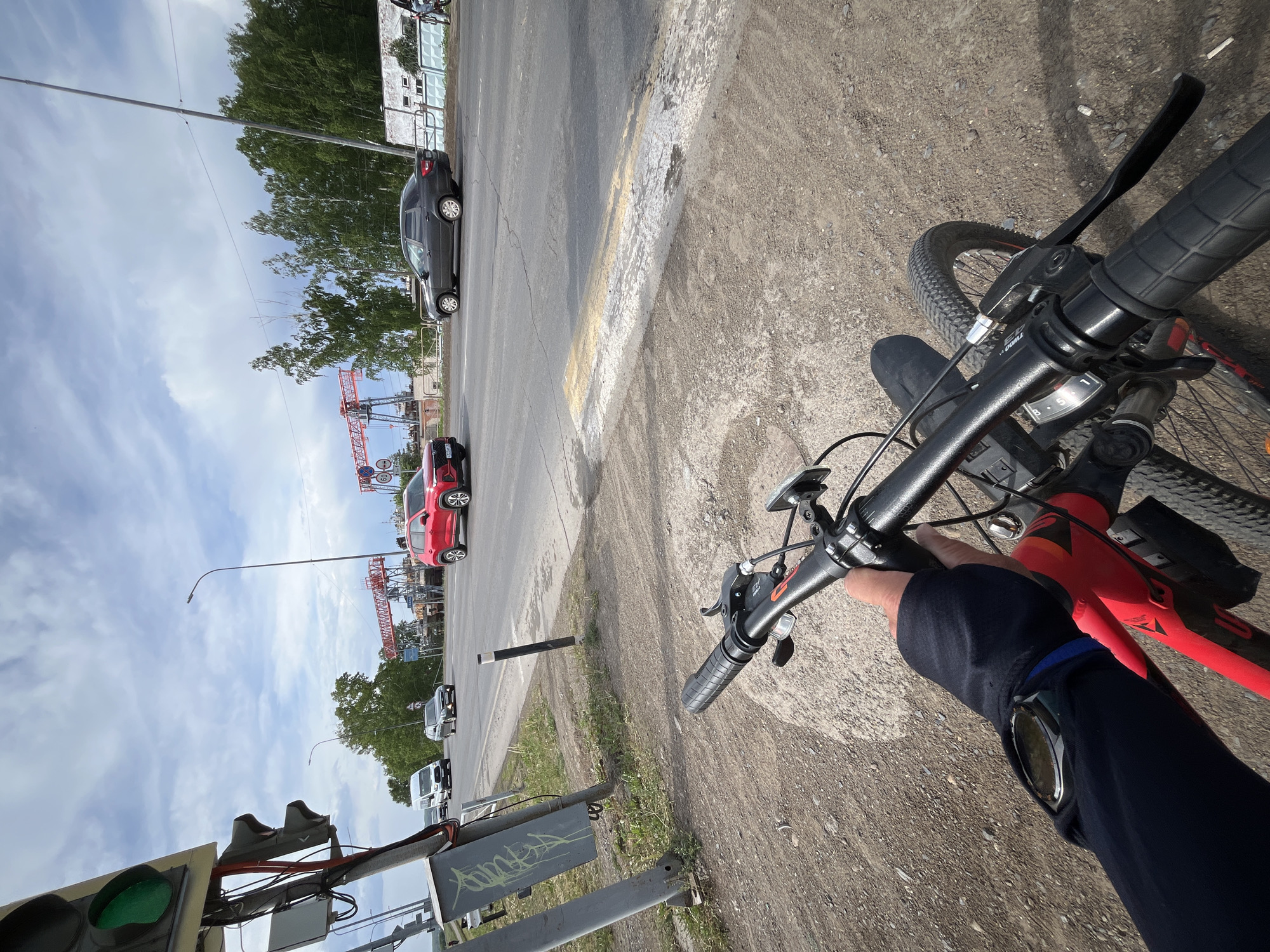 A first-person perspective photograph shows a person on a red mountain bike at a city intersection, identifiable as Chaikovsky, Russia, based on the red construction crane and architecture visible in the background. The cyclist is holding the handlebars, with the right arm visible, wearing a dark-colored sleeve and a wristwatch. The traffic light shows green, indicating permission to proceed. A gray car and a red car are stopped on the intersecting street. The location appears to be near an industrial or commercial area due to the crane and construction in the distance. The road is paved, and the shoulder is gravel. The sky is partly cloudy.