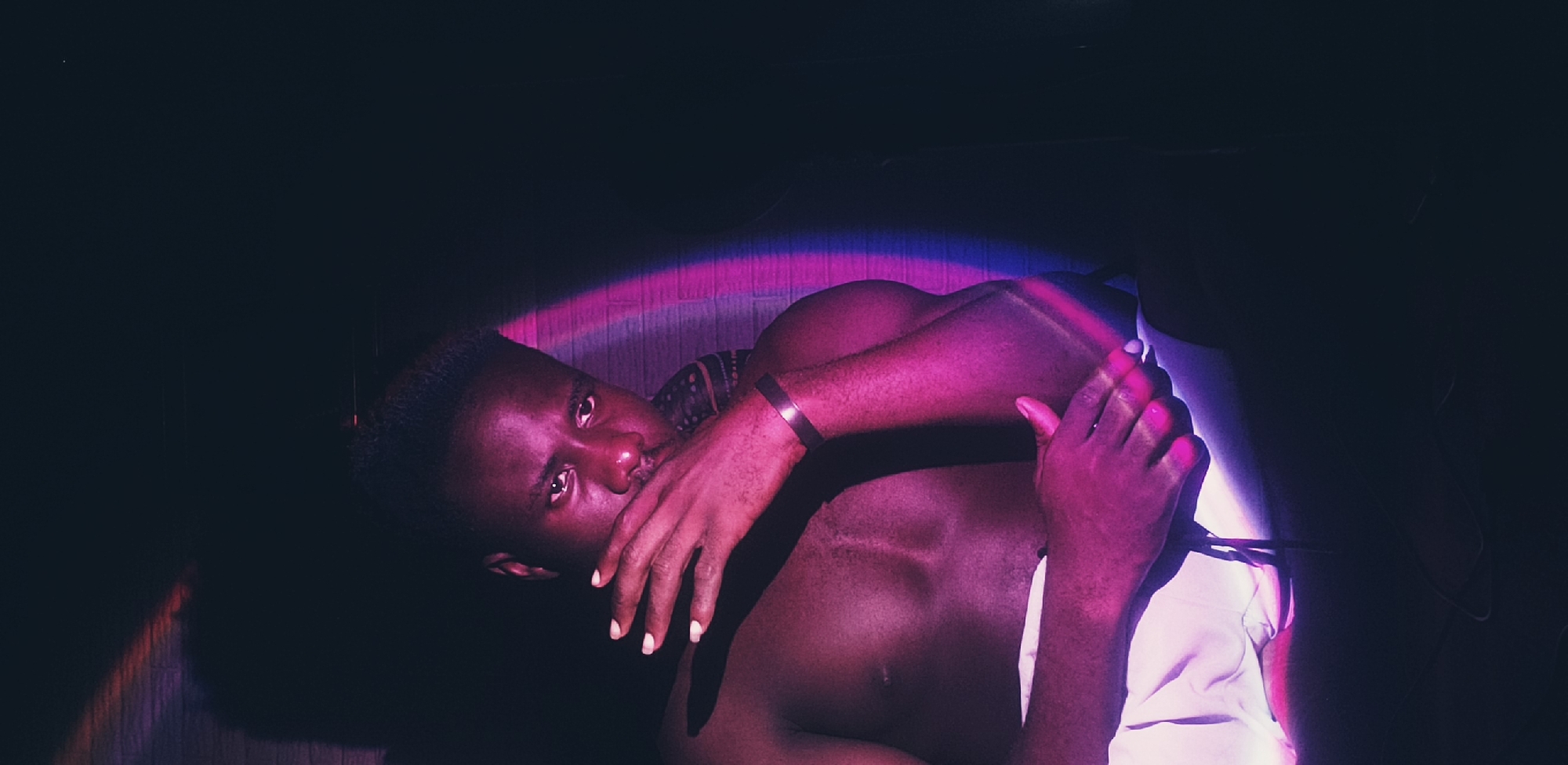 In the city of Narayi, Nigeria, a shirtless man of African descent is depicted in a dimly lit room. The man is seated and leaning forward, with his right hand covering his mouth and nose. His gaze is directed towards the viewer. He wears white shorts. The scene is illuminated by streaks of pink and purple light, creating strong shadows and highlighting certain areas of his body and face. The background appears to be a brick wall, partially visible behind him, with a dark, unlit area to the right.