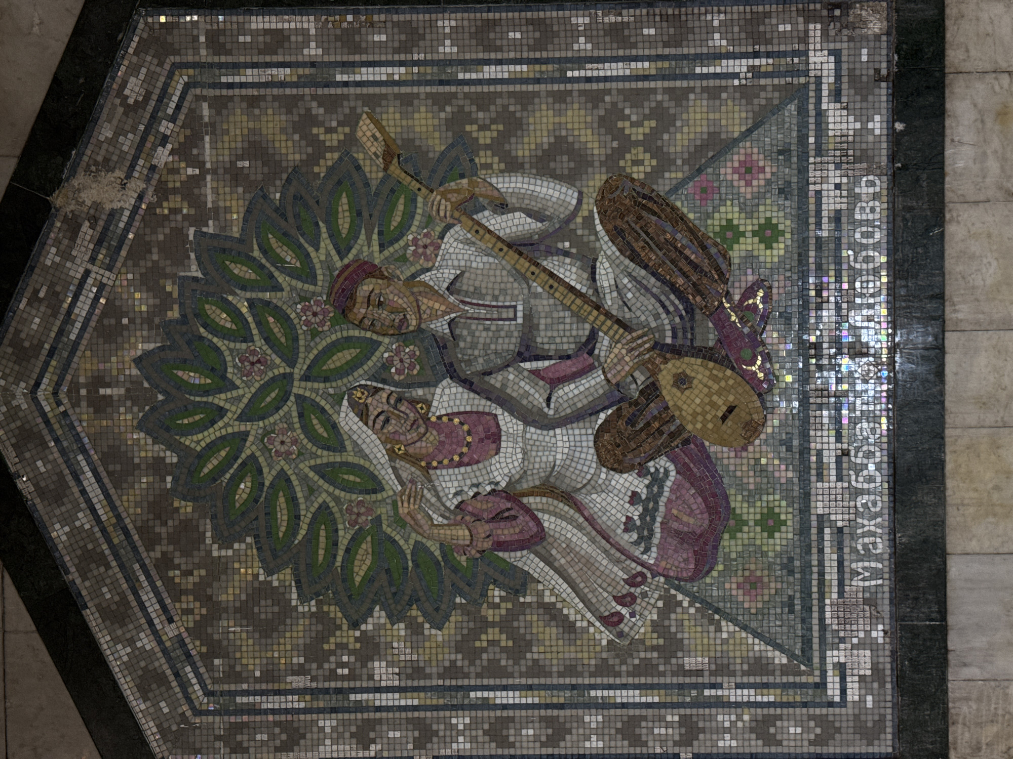 The image shows a mosaic floor panel in the Kazan Kremlin Metro station in Kazan, Russia. The mosaic depicts a man and a woman lying down. The man is playing a long-necked stringed instrument, possibly a dutar. They are surrounded by a stylized floral and leaf design. The mosaic is composed of small colored tiles. The name "Mahabbat doston" is written in Cyrillic along one edge of the mosaic. The panel is bordered by a geometric pattern of tiles and dark green stone.