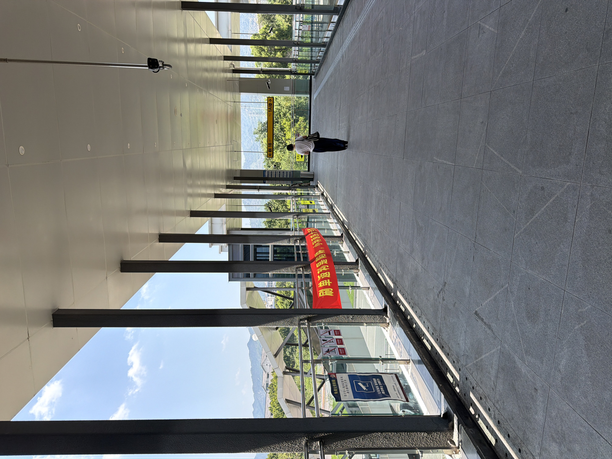An elevated, covered walkway is depicted under a bright, sunny sky. The walkway has a gray tiled floor and is supported by regularly spaced dark gray pillars. On the left side, there is a glass railing and various signs, including a blue and white sign with an eye icon. A long red banner with yellow Chinese characters is draped across the railing. In the distance, a person wearing a white shirt and dark pants walks away from the camera. The walkway overlooks a green landscape with trees and distant mountains. Overhead, there are lights and what appears to be a security camera mounted on a long pole. The walkway likely leads to or from a public transportation hub. The scene is set in Jiangjin, China.