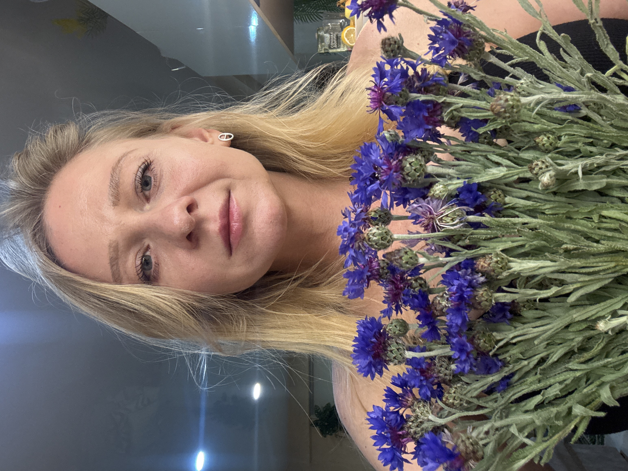 A photograph depicts a woman with blonde hair, holding a bouquet of blue cornflowers close to her face. The woman is facing the camera with a gentle smile. The background is out of focus, but elements of an interior space can be discerned. The location is within the city of Unknown, Unknown.