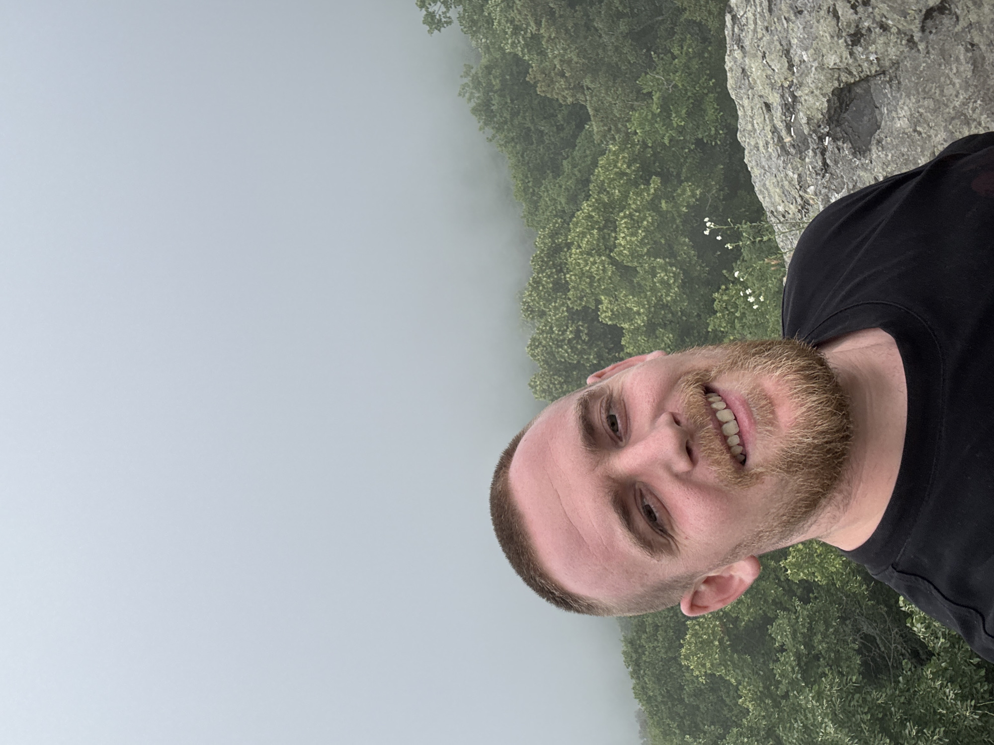 A man with light skin, short hair, and a reddish-brown beard and mustache smiles broadly at the camera, revealing his teeth. He is wearing a black t-shirt. The image is taken as a close-up, likely a selfie, giving the impression of an outdoor setting.
The background is dominated by a thick layer of light gray mist or fog that obscures the distant horizon. To the man's right, dense green trees emerge from the mist, indicating a forested area, possibly on a slope or below the vantage point. Above the trees and behind the man's right shoulder, a rugged, gray rocky outcrop or cliff face is partially visible.
The scene suggests a visit to a natural, elevated location, possibly a mountain or a coastal cliff in Sozopol, Bulgaria, during overcast or foggy weather. The man appears happy or content in the misty environment. No text is visible in the image.