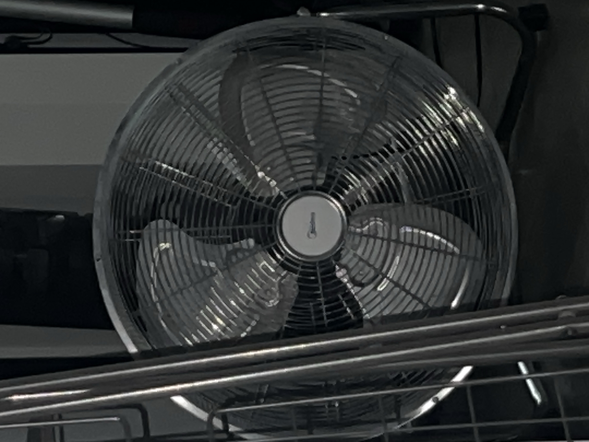 A large, circular floor fan with a metallic grille and three visible blades is positioned on the floor. The fan is silver and has a circular central hub with a logo. To the left of the fan, a portion of a silver metal rack or stand is visible, with vertical and horizontal bars. The background is dark and indistinct, with some vertical elements that could be furniture or structural components. The lighting in the image is dim, with highlights on the fan's grille and blades, suggesting a light source from the front. There are no people or animals present in the image. The scene appears to be indoors, in a dimly lit room. The image does not contain any information that would allow for the determination of a specific location in Moscow, Russia, or any other city.