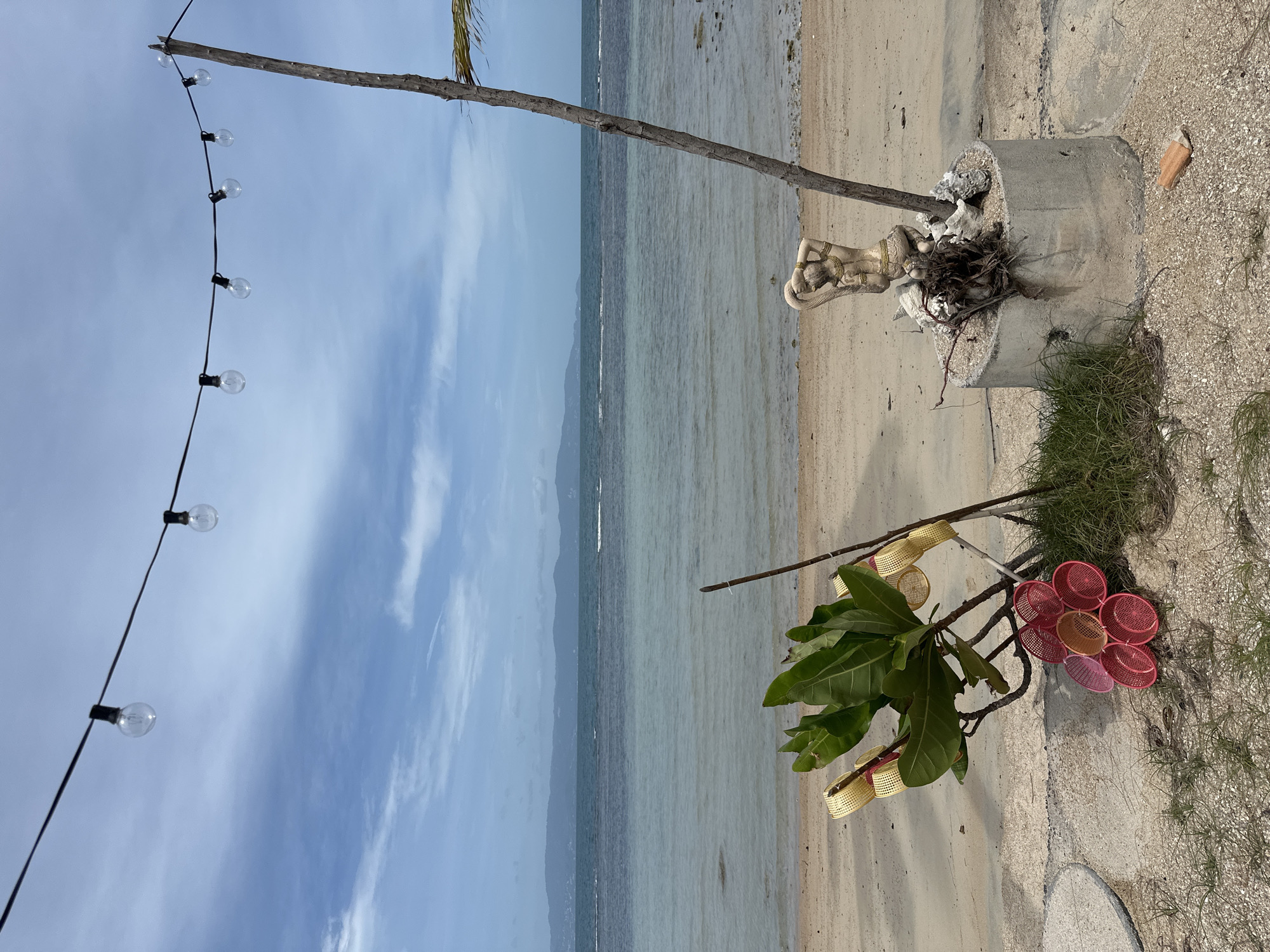A scenic beach in Ko Pha-ngan District, Thailand, features a tranquil view of the ocean under a partly cloudy sky. In the foreground, a cluster of red and pink plastic baskets are arranged on the sandy ground near a green-leafed plant. Adjacent to the plant, a concrete cylindrical structure stands with a small statue of a woman adorned with gold jewelry, positioned on top amidst shells and debris. A thin, weathered wooden pole extends upward from behind the statue. Above, a string of exposed light bulbs hangs horizontally across the frame, adding a decorative element to the outdoor setting. The beach itself is composed of sand with scattered pebbles and dry vegetation. The ocean stretches out to the horizon, where hazy mountains are visible.