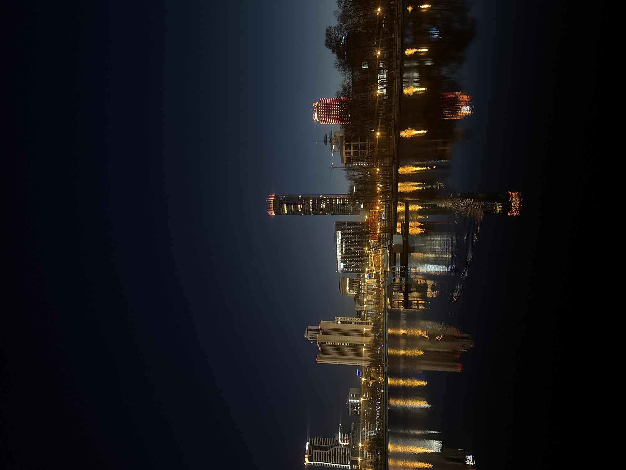 The image depicts a night scene in Yekaterinburg, Russia, featuring a cityscape reflected in a body of water. The city skyline includes illuminated buildings, notably the Iset Tower and the Vysotsky skyscraper, both distinctive architectural landmarks in Yekaterinburg. These structures are prominently lit with warm, reddish-orange lights. The reflection in the water mirrors the skyline, creating a symmetrical effect. The sky above is a dark blue, contrasting with the bright lights of the city. The scene is captured at night, with the dark environment highlighting the illumination of the buildings and their reflection. There are trees along the water edge. The overall composition suggests a calm and quiet urban scene at night.