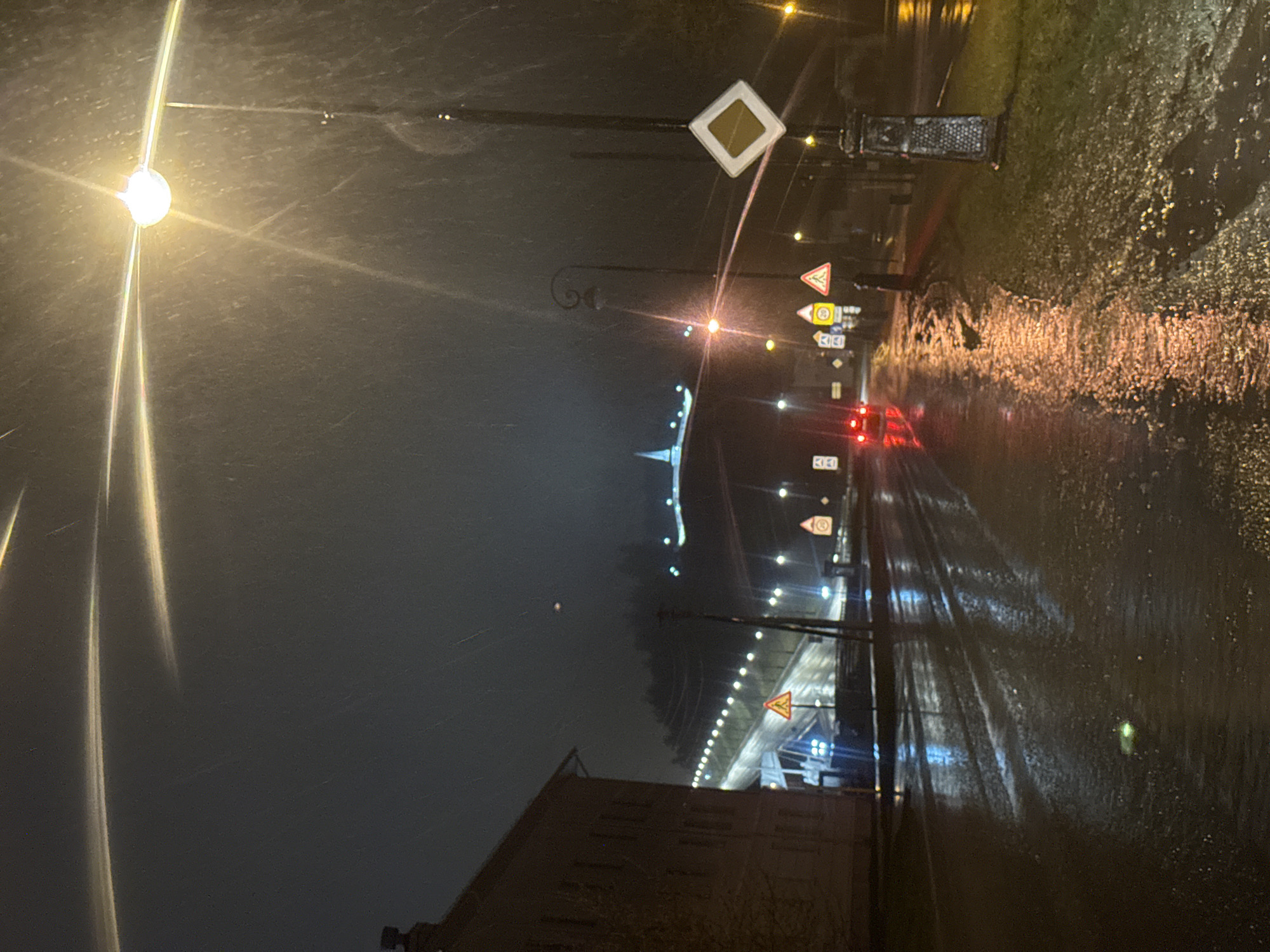 The image captures a night street scene, likely in an urban or suburban setting, under rainy or post-rain conditions.
**Subjects**:
The scene features a wet, reflective road stretching into the distance, bordered by equally wet ground or grass on its right side. Several bright streetlights illuminate the area, creating distinct lens flares, with a particularly prominent light source in the upper left. Along the road, numerous traffic signs are visible, including a prominent main road sign (a white diamond with a yellow square) and several triangular warning signs. Further down the road, the red taillights of a vehicle can be seen. In the lower left, the roofline and dark facade of a building are present. A structure with bright, decorative lighting is visible in the distant background.
**Activity or event taking place**:
The image depicts a moment on a street at night, characterized by the shimmering reflections of lights on wet surfaces, suggesting recent or ongoing rainfall. The visible vehicle taillights indicate the presence of traffic, contributing to a typical nocturnal street ambiance.
**Notable details**:
The time of day is night, with artificial lights being the primary source of illumination. The weather is wet, evidenced by the highly reflective road and ground, which mirror the lights and create a dynamic interplay of light and shadow. The bright streetlights generate strong lens flares, adding to the visual effect. The location context for this scene is Tobolsk, Russia. The overall atmosphere is moody and somewhat tranquil, despite the urban setting.
**Visible text**:
A circular speed limit sign displaying the number "30" is clearly visible. The main road sign, a white diamond with a yellow square, is also distinct. Other signs are present but their specific text or symbols are not legible.