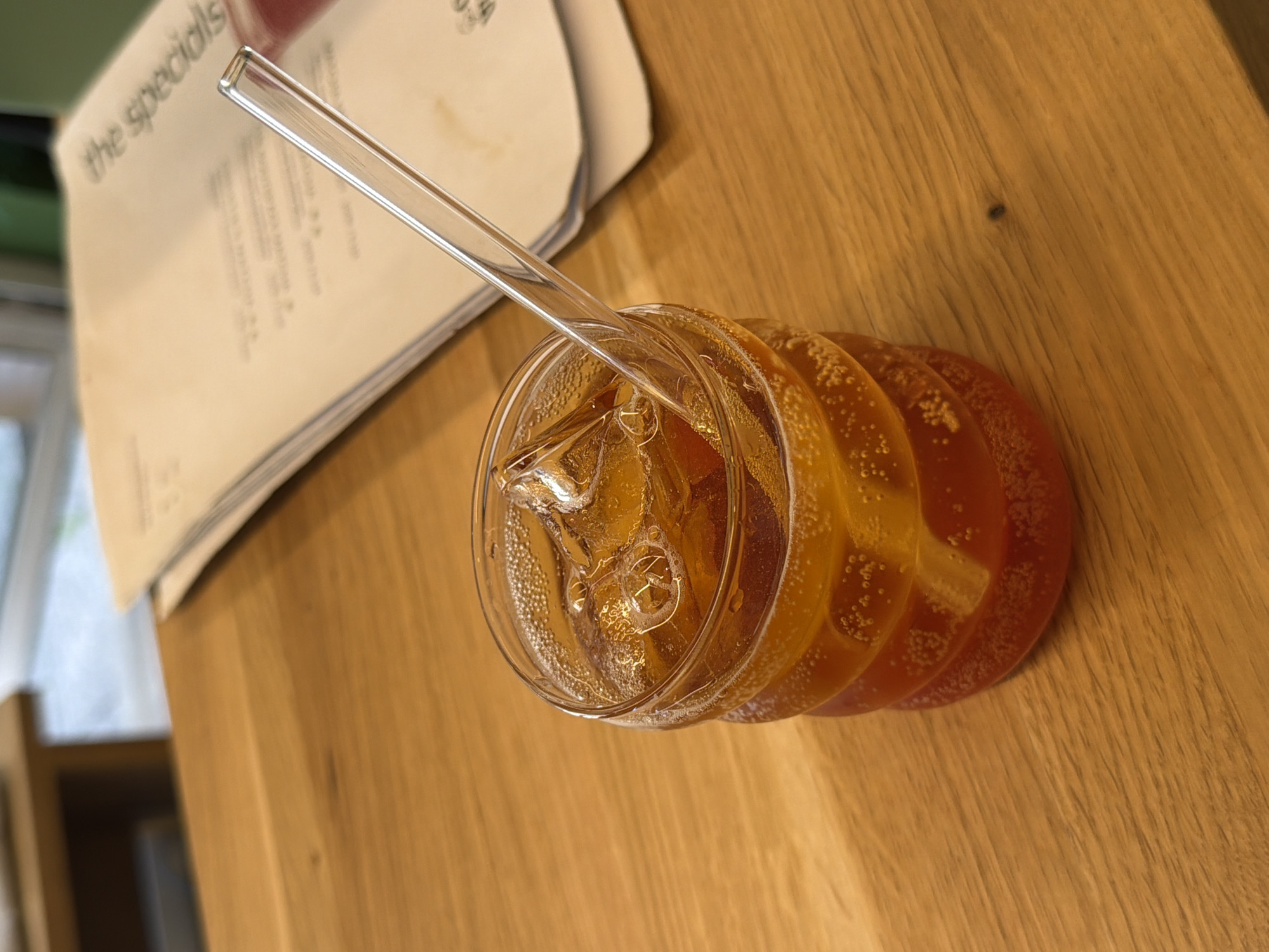 A glass of amber-colored soda with ice and a clear straw sits on a wooden table. In the background, a stack of papers with text is visible. The lighting suggests it is daytime. The scene is likely indoors, possibly in a cafe or restaurant. There is no indication of the location being in Sofia, Bulgaria.