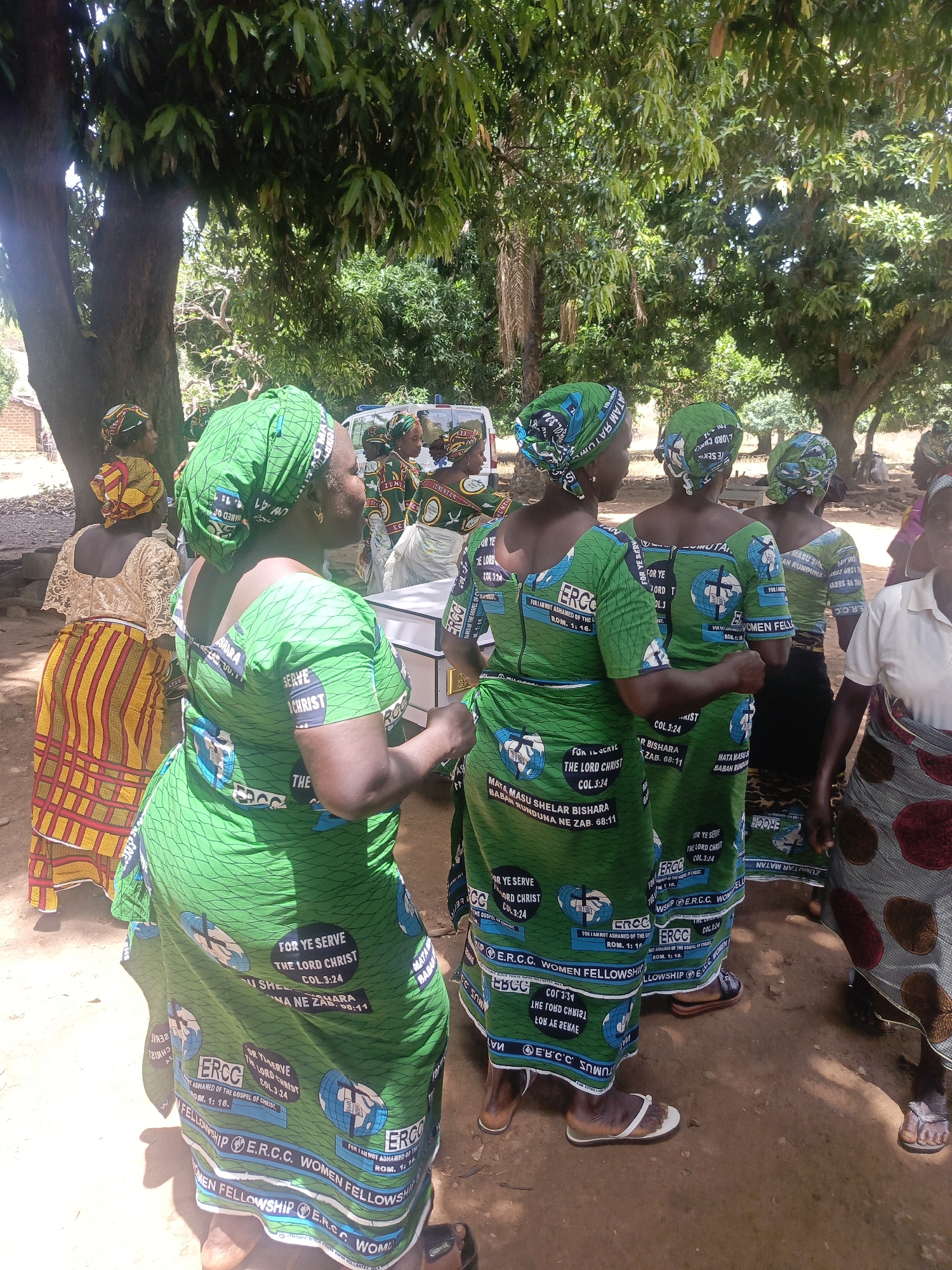 A group of women stand together in an outdoor area, likely in Ninzo South, Nigeria. The women, many dressed in matching green outfits, with the inscription "E.R.C.C. WOMEN FELLOWSHIP" and the phrase "FOR YE SERVE THE LORD CHRIST COL.3:24" printed on them. They are under the shade of trees. A few women are wearing different patterns. A white ambulance can be seen in the background. The ground appears to be dirt.