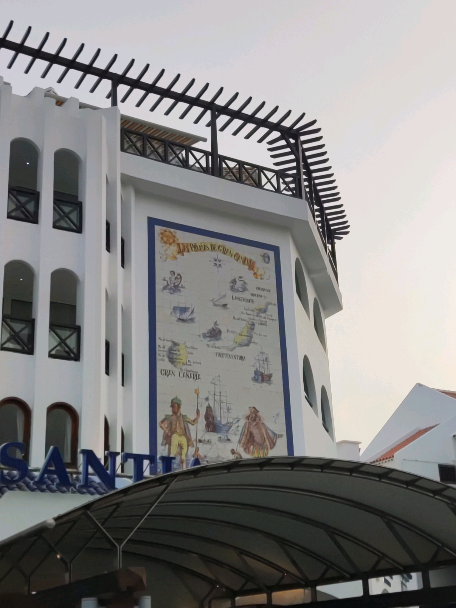 The image shows a white building with arched windows and balconies. A large mural depicting a map and figures is on the side of the building. The mural features a sun, ships, and people in historical attire. Text on the mural is in French and Spanish and includes the words "LES PAYS DE GRAND CANARIA" and "GRAN CANARIA". The building is partially obscured by a dark, modern canopy structure in the foreground. The sky is overcast. The location is identified as Playa De Las Américas, Spain, suggesting this is a hotel or similar establishment.