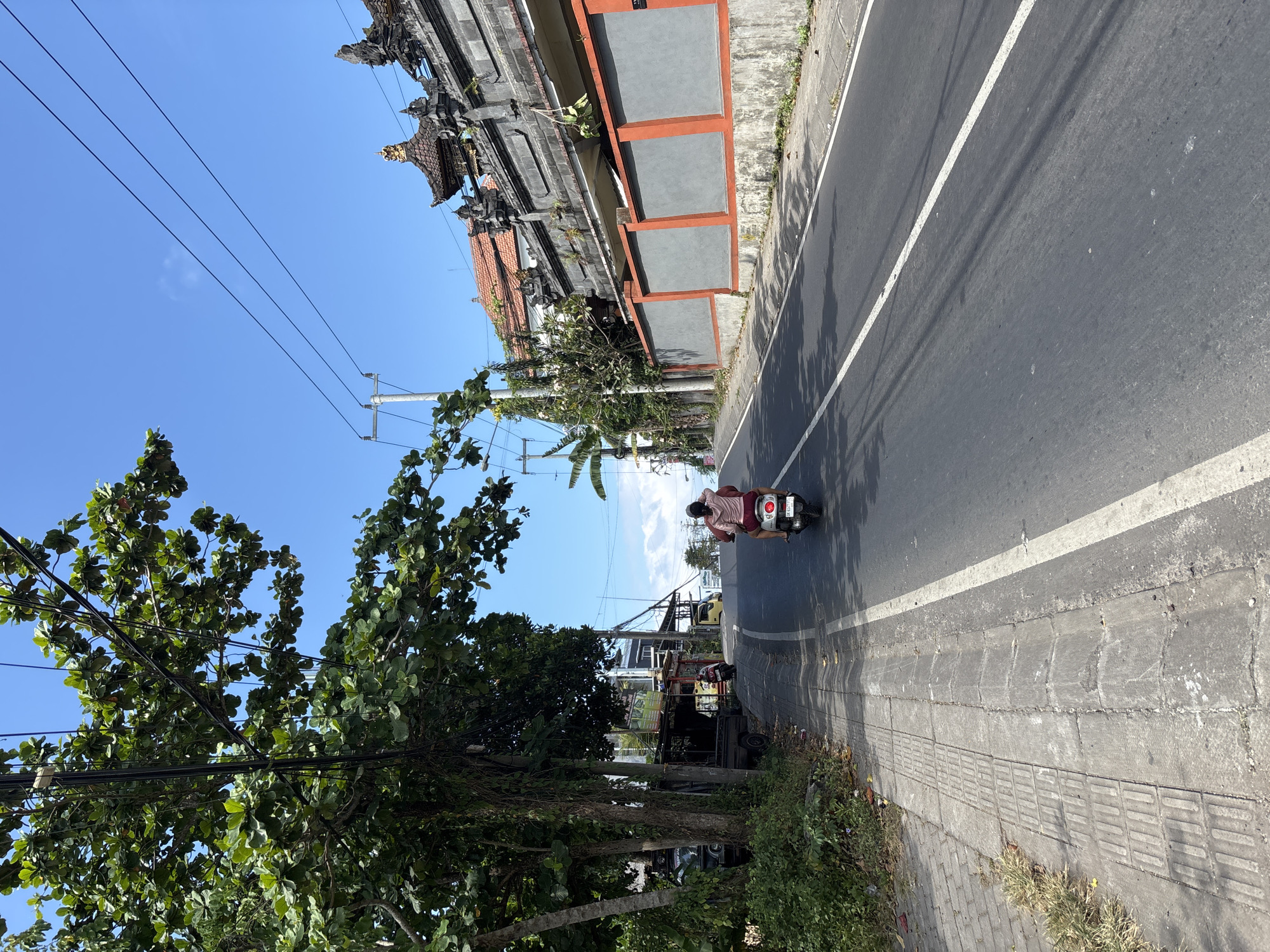 This bright, sunlit image depicts a street scene in what appears to be a tropical urban or semi-urban environment, consistent with Kuta Selatan, Indonesia. The photograph is taken from a slightly elevated perspective, looking down a paved asphalt road that curves gently into the distance.
In the center of the road, a person wearing a light-colored top is riding a white scooter away from the viewer. The scooter's license plate is clearly visible as "DK 6479 QAB". The road has a distinct white solid line marking the center or edge, and strong shadows are cast across its surface, suggesting bright mid-day sun.
To the left of the road, lush green trees with dense foliage partially frame the scene. Power lines crisscross through the branches and against the clear blue sky. A sidewalk with textured concrete pavers runs along the left side.
On the right side of the road, a detailed sidewalk with patterned concrete abuts a low wall with grey panels set into orange-brown frames. Beyond this, a prominent building features traditional Balinese architecture, characterized by an ornate dark grey stone facade with intricate carvings and a multi-tiered roof topped with golden accents. Further down the street, other structures and commercial signs are visible, hinting at an active area. The sky is a cloudless, vibrant blue.