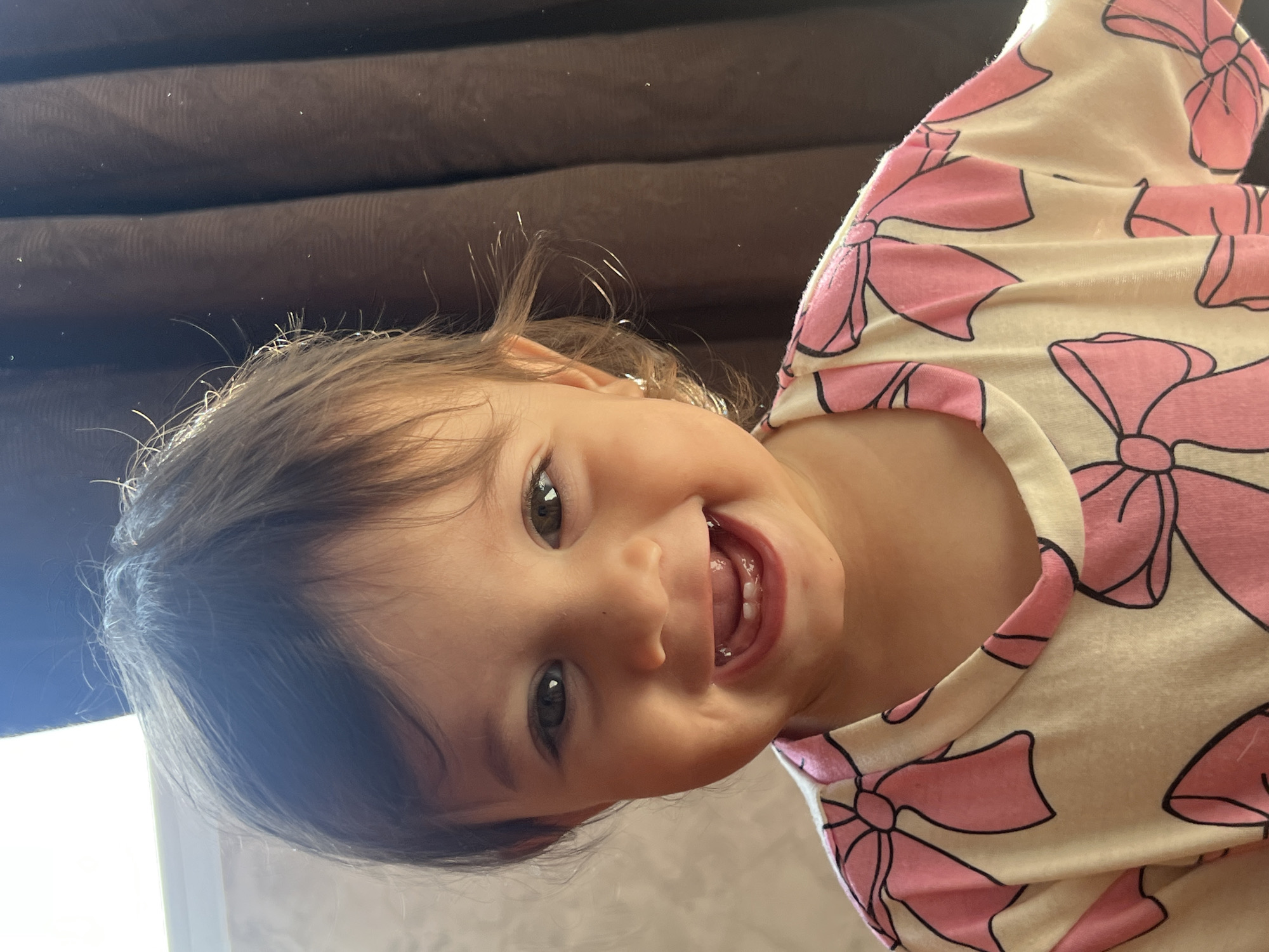 A close-up, eye-level shot shows a young toddler with brown hair and brown eyes smiling broadly, revealing several teeth. The child is wearing a light-colored shirt with a pattern of pink bows. The background is out of focus, with a textured wall on the left and a dark brown curtain on the right. Sunlight casts a bright glow on the left side of the child's face and hair. The image appears to have been taken indoors, possibly in a residential setting. There is no discernible information in the image that would indicate a specific location in the city of Elena, Bulgaria.