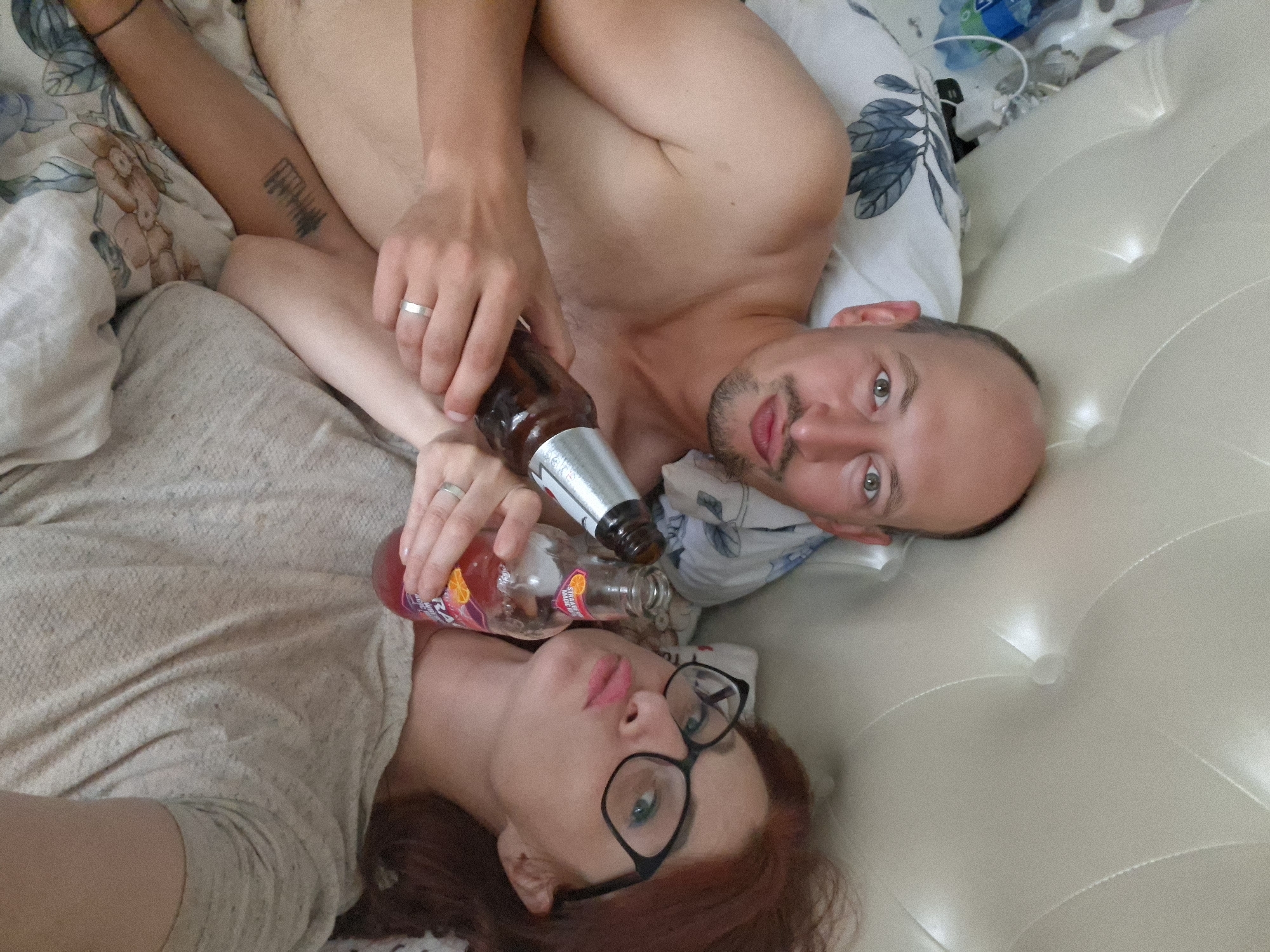 In a bed, a man and a woman lie down, each holding a bottle. The man, bald with a goatee, looks at the camera. The woman, with red hair and glasses, also faces the camera. Both have their lips pursed. The man's left arm is raised, holding a bottle of what appears to be an alcoholic beverage, while the woman holds a bottle of a pink beverage. The setting appears to be a bedroom in Myadzyel, Belarus.