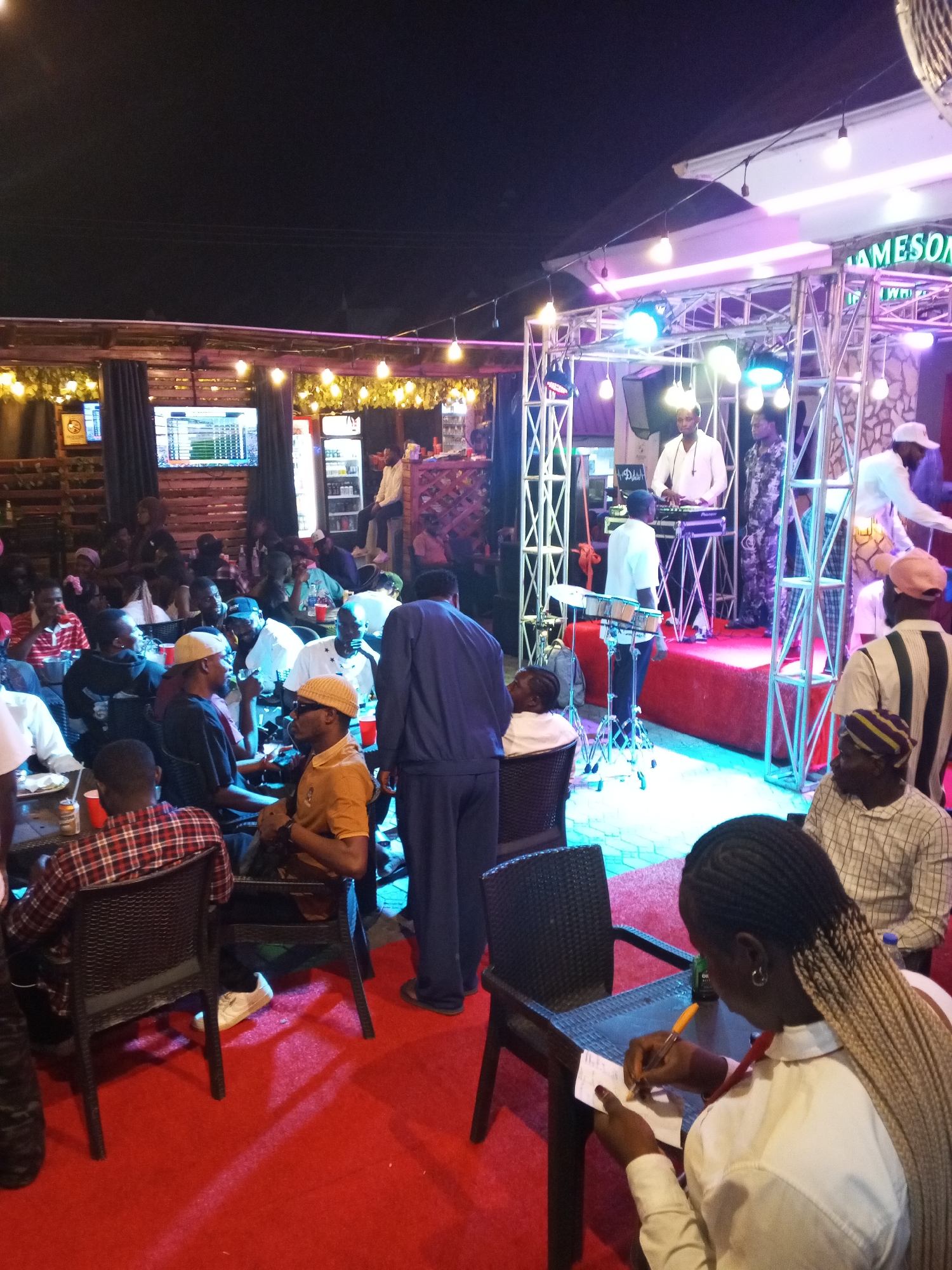The image depicts a lively outdoor event at night, likely a performance or gathering, taking place in Lokoja, Nigeria. A stage with bright, colorful lights is set up, featuring musical equipment such as a DJ console and a drum set. Several performers are visible on the stage, some interacting with the equipment. A crowd of people is seated at tables and chairs, watching the performance. The environment appears to be a restaurant or bar with a red carpeted floor and outdoor seating. The overall atmosphere is social and festive.