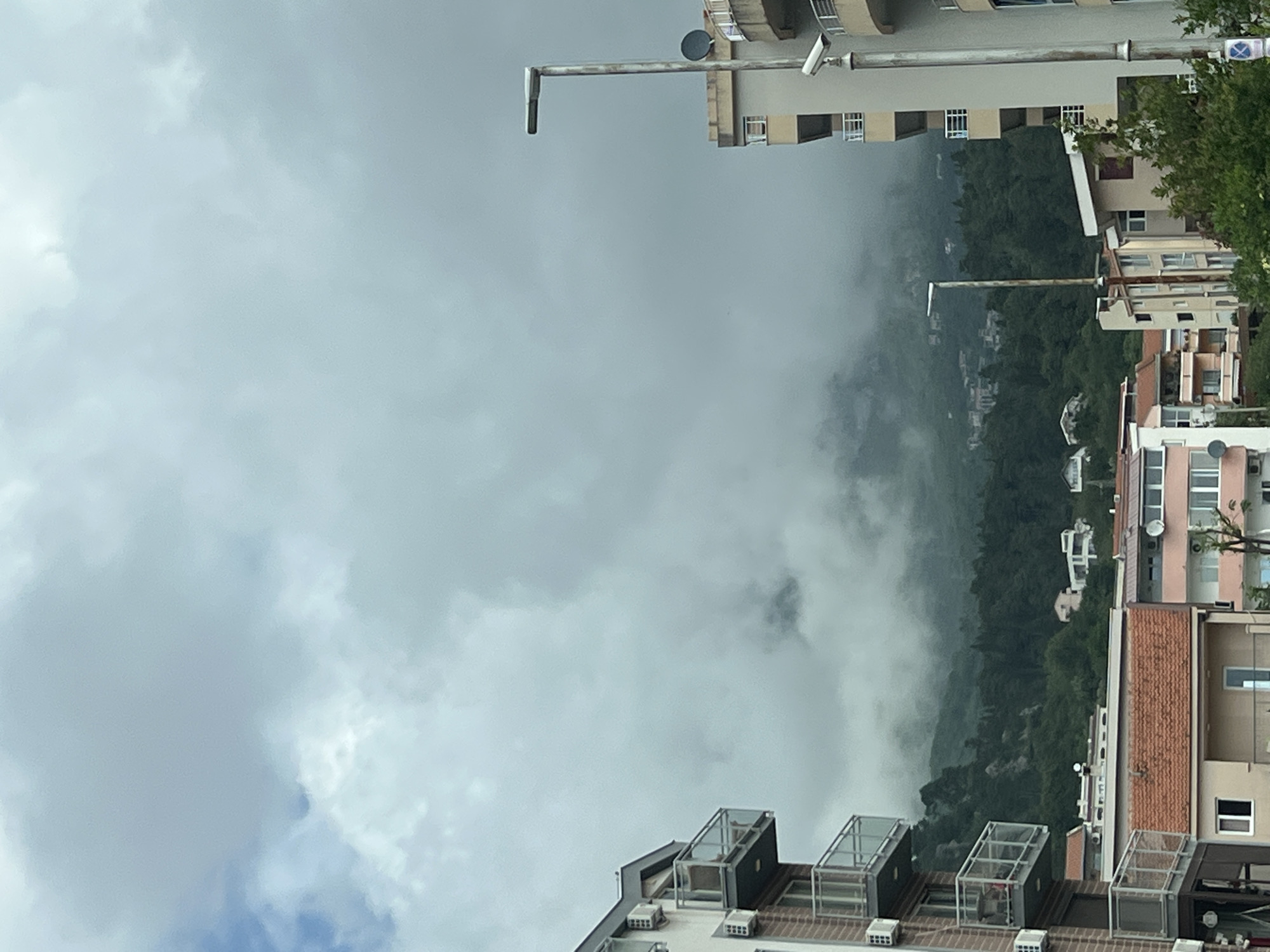 The image depicts a view in Budva, Montenegro, characterized by a cloudy sky covering a significant portion of the frame. Below the clouds, a mountainous landscape is visible, partially obscured by fog or low-lying clouds. Interspersed on the mountainside are residential buildings.
The lower section of the frame features the rooftops of several apartment buildings. These buildings have distinctive architectural elements such as enclosed rooftop structures. Above and to the left, the image shows a building with an antennae on top.