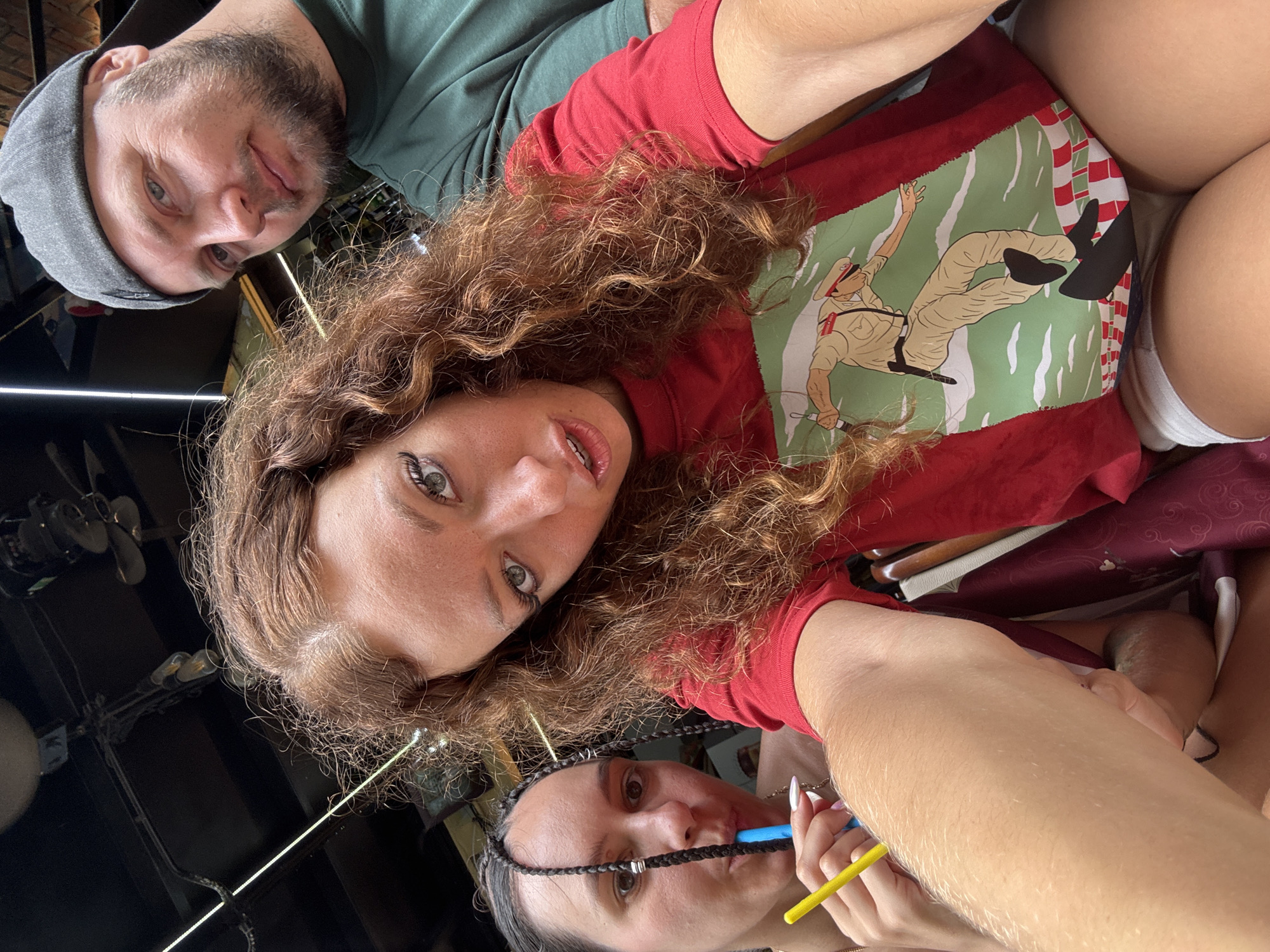 Three people are visible in the photograph. A man with a beard and a gray cap is on the left side of the image. A woman with long, curly brown hair is in the center, wearing a red shirt with a cartoon graphic. Another woman, who is braided, is in the bottom part of the image. She is holding a straw in her mouth. The environment around them suggests an indoor setting, potentially a restaurant or cafe, with industrial-style lighting and fans visible on the ceiling. The location is noted as Hà Nội, Vietnam.