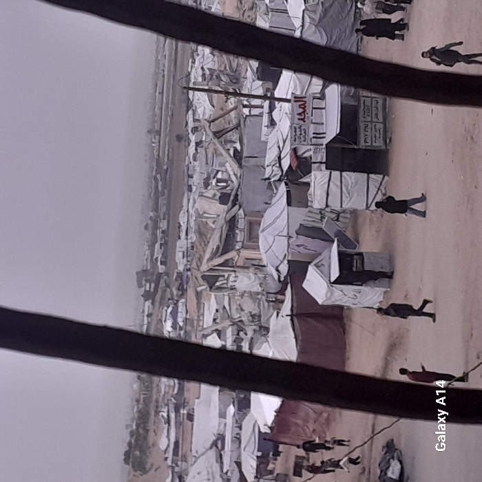 The image shows an overcrowded refugee camp with makeshift shelters. White tents are densely packed together, some appearing damaged. A dusty, barren landscape surrounds the camp. Several people are visible, walking through the camp, their figures small against the vastness of the scene. The photo appears to have been taken from a high vantage point, looking down at the settlement. The image evokes a sense of displacement, poverty, and vulnerability.
