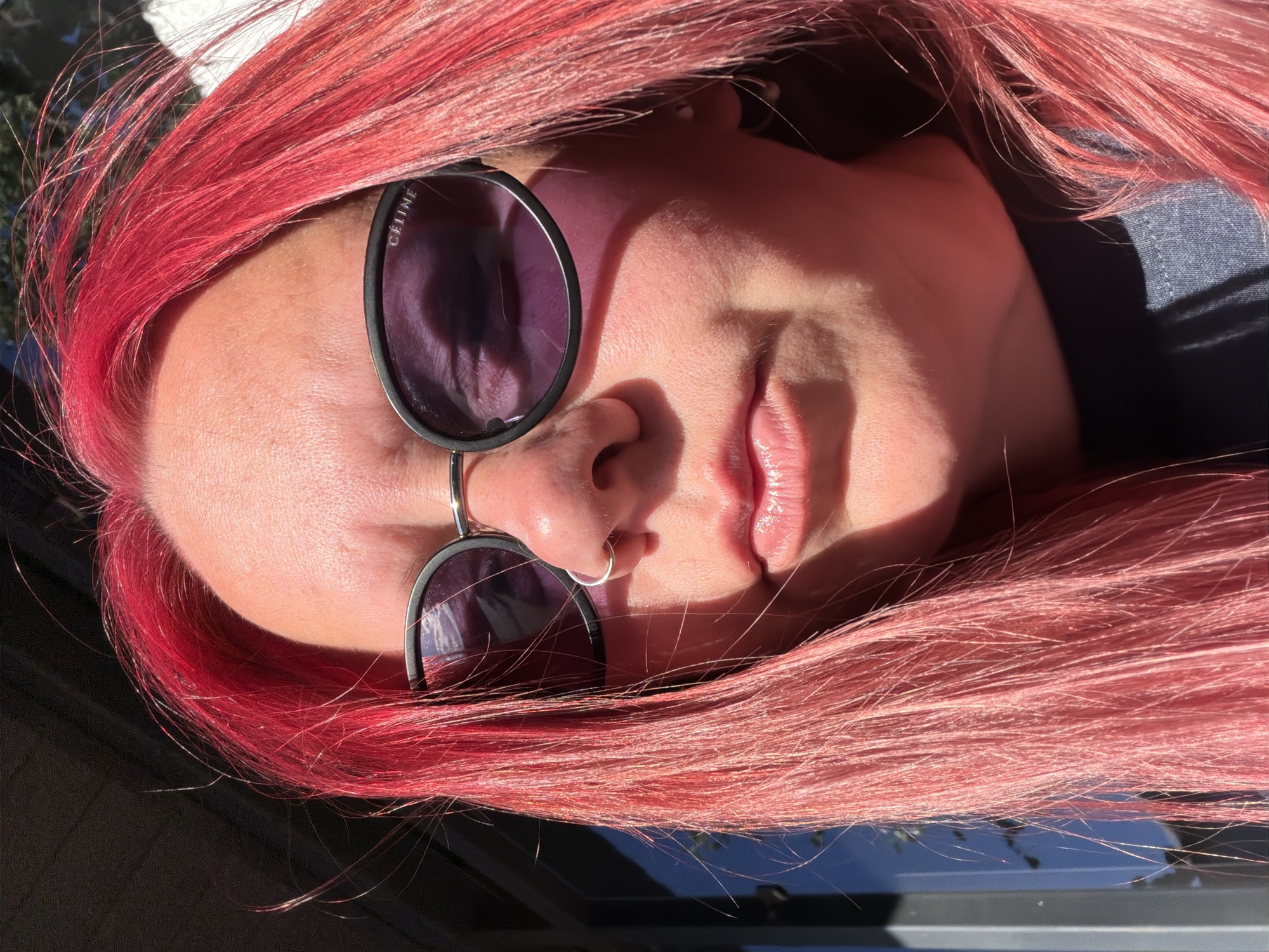 A close-up, outdoor, headshot of a woman with bright pink hair, wearing sunglasses with black frames and the brand name "CELINE" visible on the side. She has a silver nose ring and a small silver earring. The lighting suggests it is daytime, with strong sunlight creating shadows on her face and hair. The background is blurred but appears to be an urban or semi-urban setting with some vertical structures and foliage visible. There is no information about the location being Chaikovsky, Russia, or any specific city.