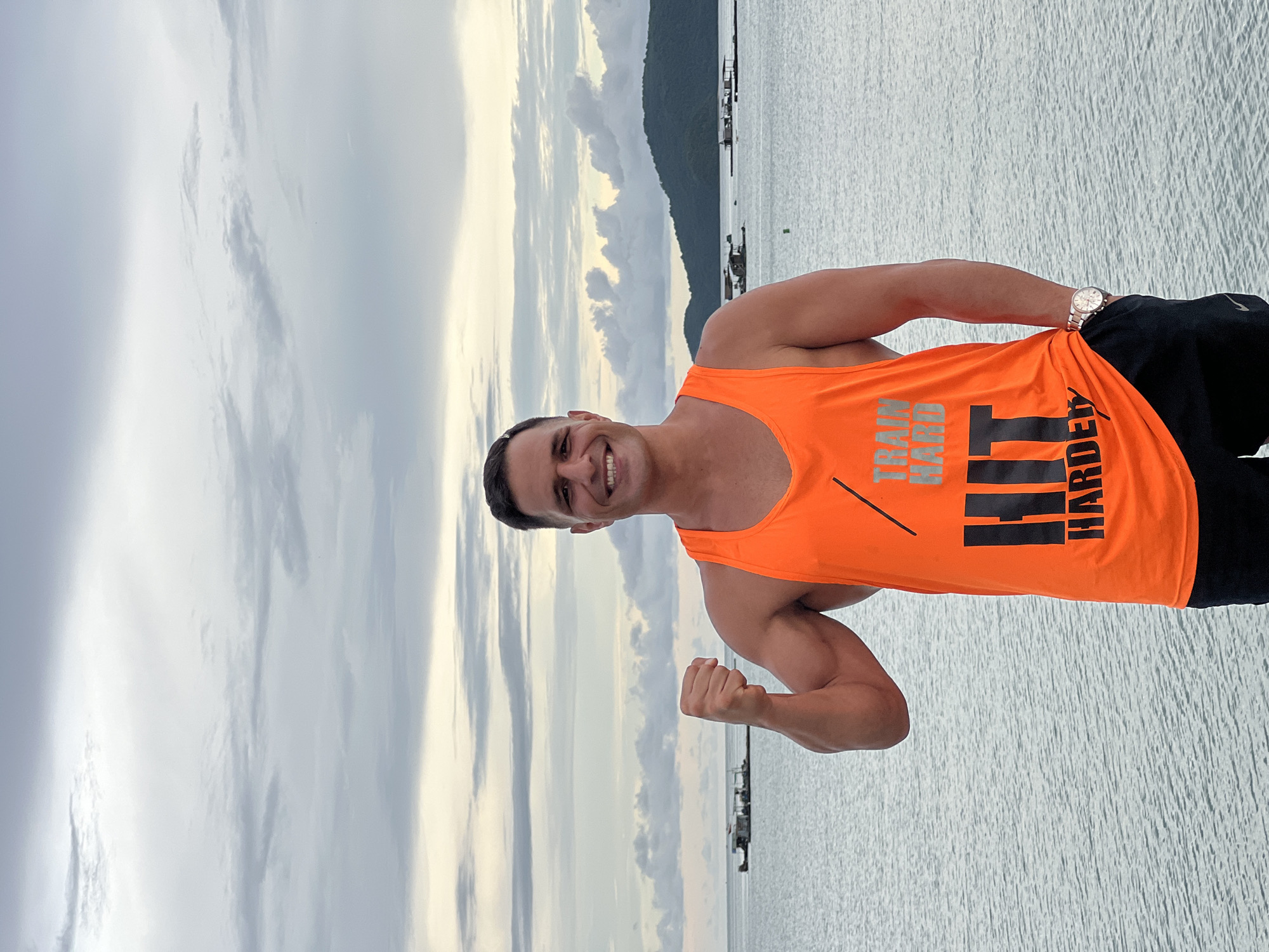 A man stands with his right arm flexed, wearing an orange tank top with the words "TRAIN HARD HIT HARDER" printed on it, and black shorts. He is smiling and looking at the camera. Behind him, the ocean stretches out to a distant, tree-covered island under a cloudy sky. Several small structures, possibly boats or fishing platforms, are visible on the water. The scene appears to be in Chalong, Thailand, given the coastal environment and tropical island in the background.