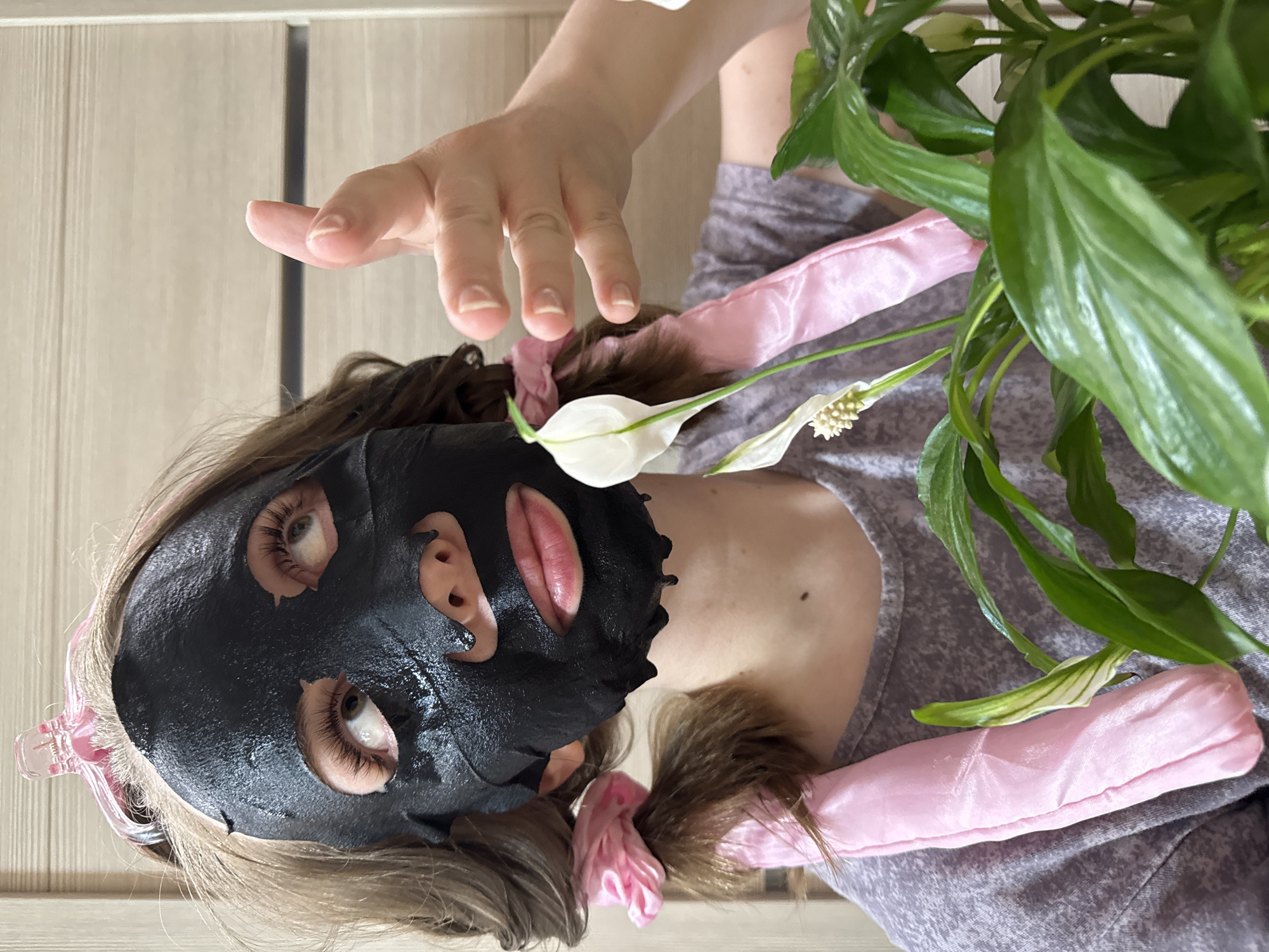 The rotated image captures a person engaged in a self-care routine within an indoor setting, likely a home. The central subject is a person, appearing to be lying down, wearing a black sheet mask that covers their entire face except for circular cutouts for their eyes and an oval opening for their mouth. Their eyes are open and visible through the mask, appearing neutral in expression, with visible eyelashes. Their lips are parted slightly.
Their brown hair is styled in two pigtails or buns, secured with fluffy pink satin scrunchies or hair ties on either side of their head. Another pink satin object, possibly a heatless hair curler, rests on their shoulder/chest area, near the base of their neck. The person is wearing a grey, textured or knitted casual top.
To the right side of the frame, a healthy green plant resembling a Peace Lily (Spathiphyllum) is prominently featured, with glossy green leaves and distinctive white, spathe-like flowers with yellow spadices. The person's right hand is extended forward from the top of the frame, fingers slightly curled, as if reaching or posing. The background consists of a light-colored, vertically paneled surface, suggesting a wooden wall or door. The lighting appears soft and even, typical of an indoor environment during daytime, with no strong shadows to indicate harsh sunlight. There is no visible text in the image. The overall mood conveyed is one of relaxation and personal grooming.