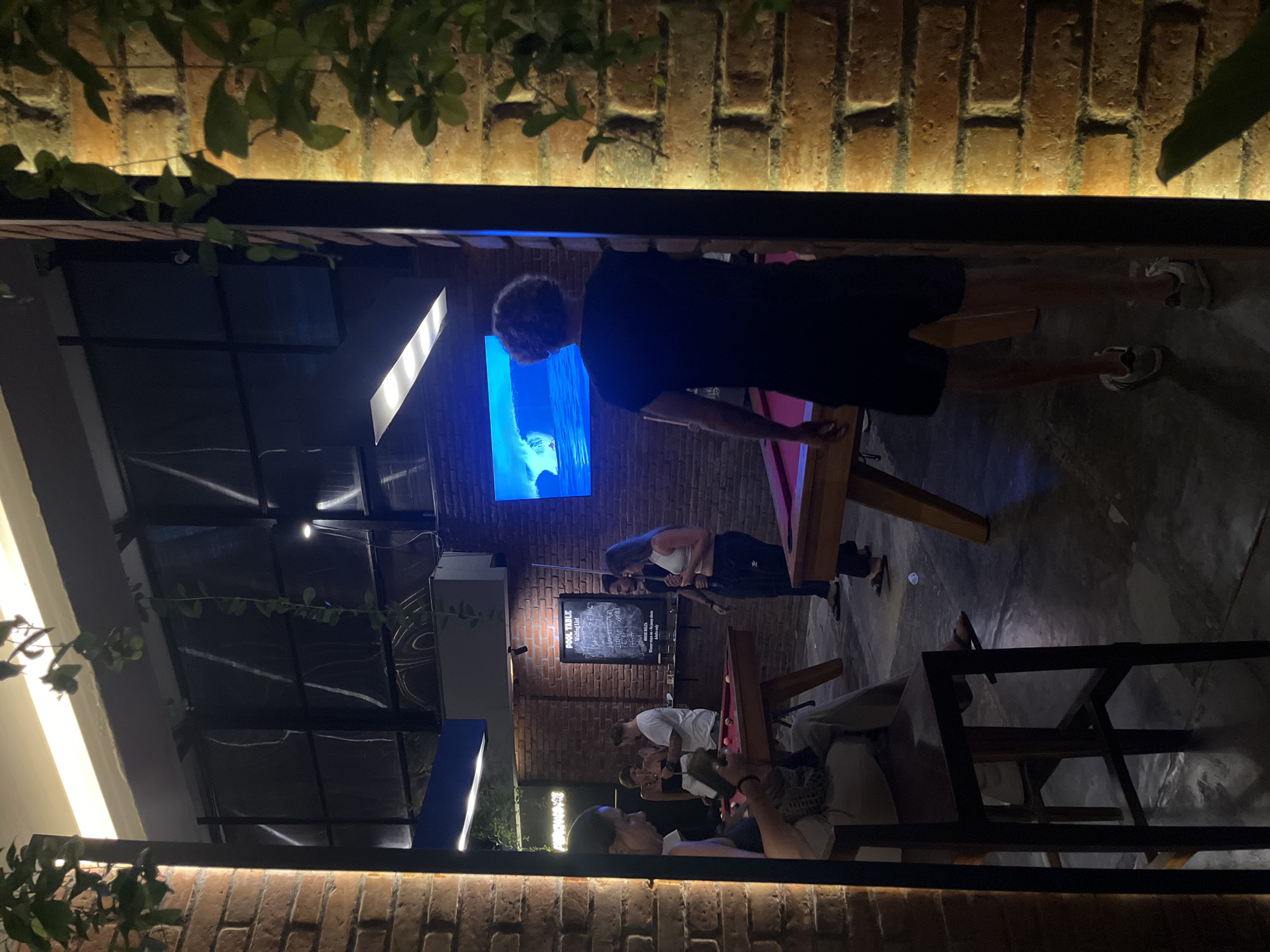 A night scene at a bar in Kuta Selatan, Indonesia. The bar has a brick wall and is decorated with plants. A blue television screen is visible on the wall. There is a pool table, and several people are playing or watching a game. One man stands at the pool table, appearing to be lining up a shot. Another group of people is seated at a table in the foreground. A chalkboard with a “Pool Table Waiting List” is also seen.
